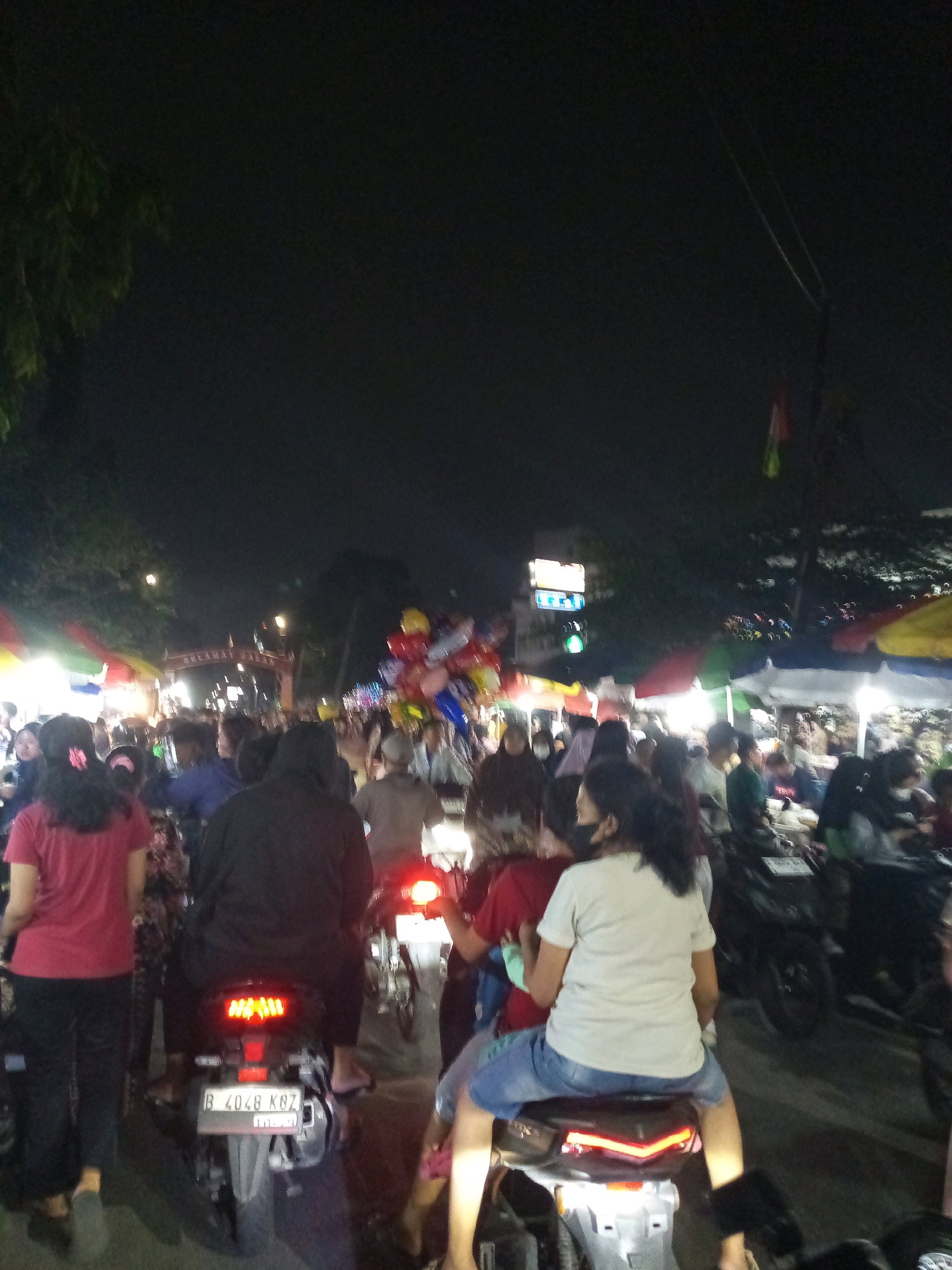 The image depicts a night scene in the city of Bekasi, Indonesia, characterized by a busy street market or event. In the foreground, several individuals are seen riding motorcycles. One motorcycle in the center has a license plate visible that reads "B 4048 KBZ". The rider of this motorcycle is a person wearing a light-colored t-shirt and blue shorts, with a dark-colored mask covering their lower face. A person in a black hooded jacket is seated behind the rider. To the left, a person in a red t-shirt and black pants is walking. In the background, there is a crowd of people, numerous market stalls with colorful umbrellas, and a lighted archway that appears to say "SELAMAT DATANG PASAR" (Welcome Market). Balloons are also visible in the background, adding to the festive atmosphere. The overall environment suggests a lively outdoor gathering at night.
