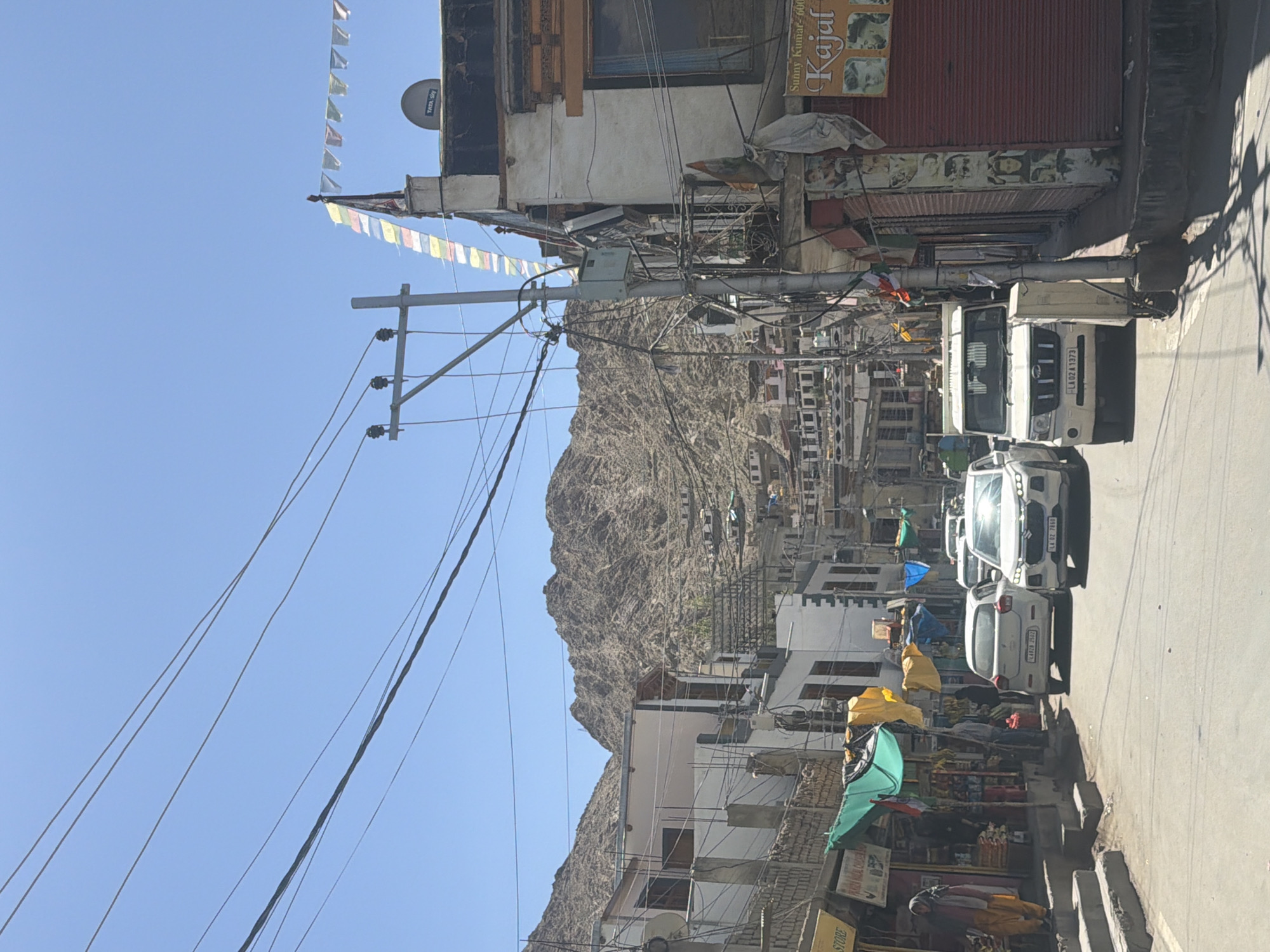 The image depicts a street scene in the city of Leh, India, characterized by a narrow road lined with buildings and a steep, rocky mountain in the background. The sky is clear and blue.
On the left side of the road, there are several shops with awnings and goods displayed. One shop has a sign that reads "STORE" and another sign advertising "Chicken". A woman in a traditional outfit is visible standing on some steps.
The road itself is paved and has several vehicles, including a white car, a white SUV, and a white Mahindra Bolero. The cars are stopped or moving slowly in traffic.
On the right side of the road, there are more buildings, including one with a sign for "Kajal". Prayer flags are strung across the scene, attached to utility poles. A satellite dish is visible on the rooftop of one of the buildings.
The overall environment suggests a bustling, urban area within a mountainous region, with a blend of commercial activity and local life. The presence of prayer flags and the architectural style of the buildings hint at a location with a significant Buddhist cultural influence. The utility poles and numerous overhead wires are a prominent feature of the urban landscape. The lighting indicates it is daytime, likely midday due to the bright sun.