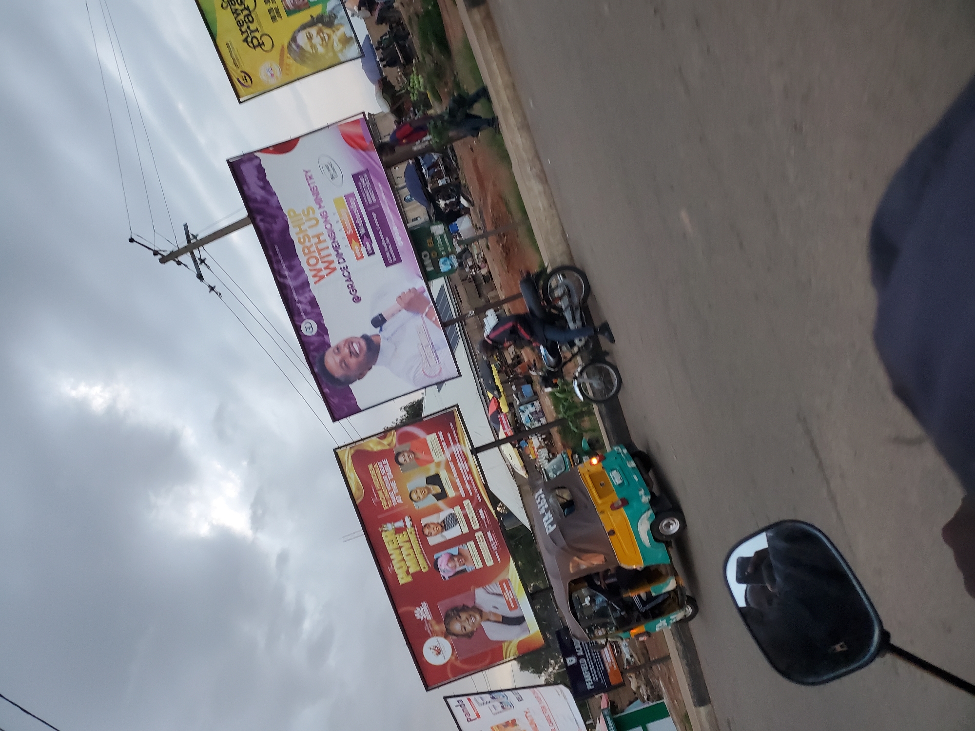 A street scene in Gura Top, Nigeria, is captured from a first-person perspective, likely from a motorcycle, as indicated by the rearview mirror reflecting the rider. The foreground shows the mirror and part of the rider's arm, which is out of focus. The main focus of the image is a street lined with various billboards and a three-wheeled vehicle, commonly known as a "Keke Napep," in motion.
The billboards are prominent and advertising different services and events. One billboard, featuring a man with a microphone, reads "WORSHIP WITH US @GRACE DIMENSIONS MINISTRY." Another, in red and gold, displays the text "POWER MOVE COMPELS" and features portraits of four women, suggesting a women's empowerment event or similar gathering. A third billboard, partially visible, is yellow and features the text "Arewa Praise."
The Keke Napep, a turquoise and yellow vehicle, is positioned in the middle ground, driving on the asphalt road. A motorcycle with a rider is also visible to the right of the Keke Napep, and another motorcycle is parked in the left middle ground. Several people are seen in the background, some walking, and others gathered around stalls, suggesting a market or commercial area. The environment appears to be a typical Nigerian urban setting with a slightly overcast sky. The overall impression is that of a busy street with vibrant advertising and local transportation.