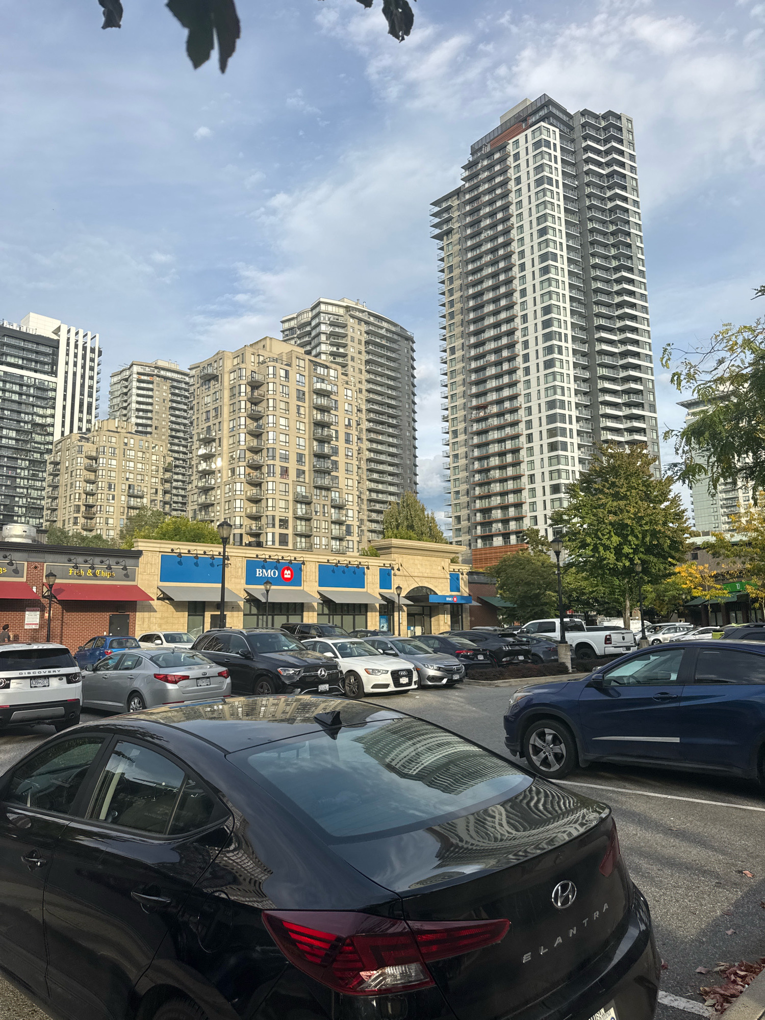 The image shows a street view in New Westminster, Canada, featuring a row of apartment buildings and a shopping area. In the foreground, several cars are parked, including a black Hyundai Elantra. Behind the parked cars, a strip of businesses is visible, with awnings and storefronts. One prominent sign reads "BMO," indicating a bank. Further back, several tall, modern apartment buildings dominate the skyline, characterized by their numerous balconies and glass windows. The sky above is partly cloudy. The scene depicts a typical urban environment with a mix of residential and commercial structures.