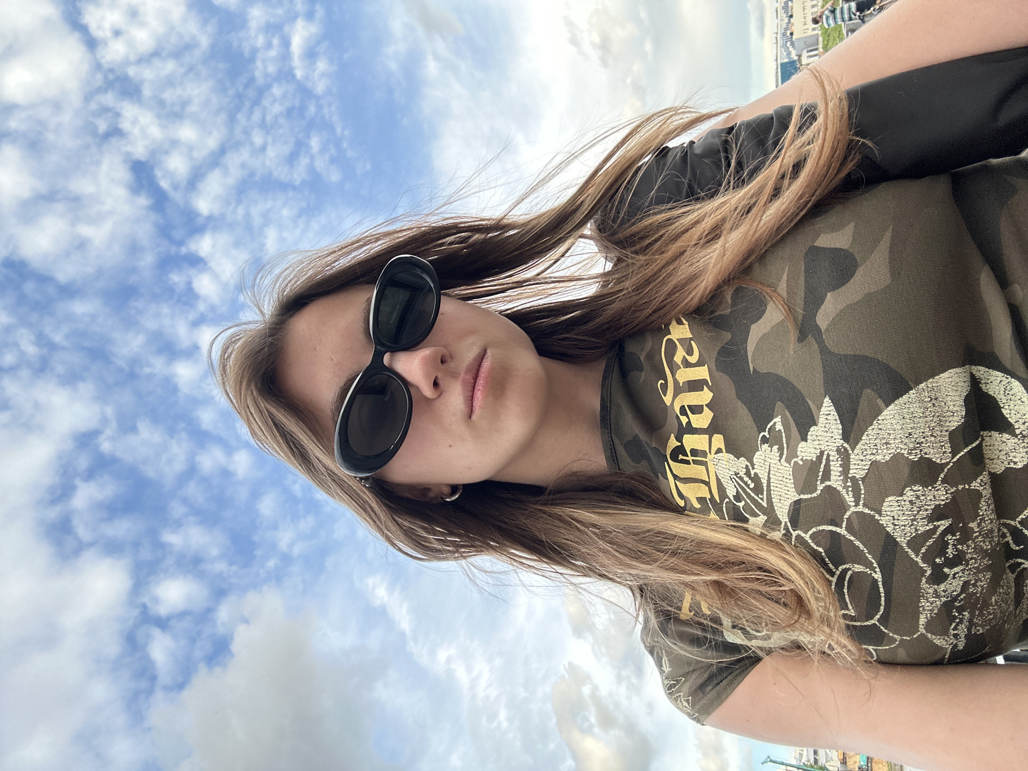 A young woman with long, light brown hair, wearing black oval sunglasses and a camouflage t-shirt with gold lettering and a floral pattern, is pictured outdoors against a blue sky filled with white clouds. She is looking directly at the camera with a neutral expression. Her hair is windswept. The t-shirt has a stylized gold text that appears to read "FARA" or "FLORA" in a gothic font, accompanied by a delicate floral design. In the background, a blurred cityscape with buildings and what appears to be a body of water or a park can be seen, suggesting an urban or park setting. The lighting indicates it is daytime, likely a clear and breezy day. The orientation of the image suggests it's a selfie taken outdoors, possibly on a balcony or elevated viewpoint given the distant city skyline.