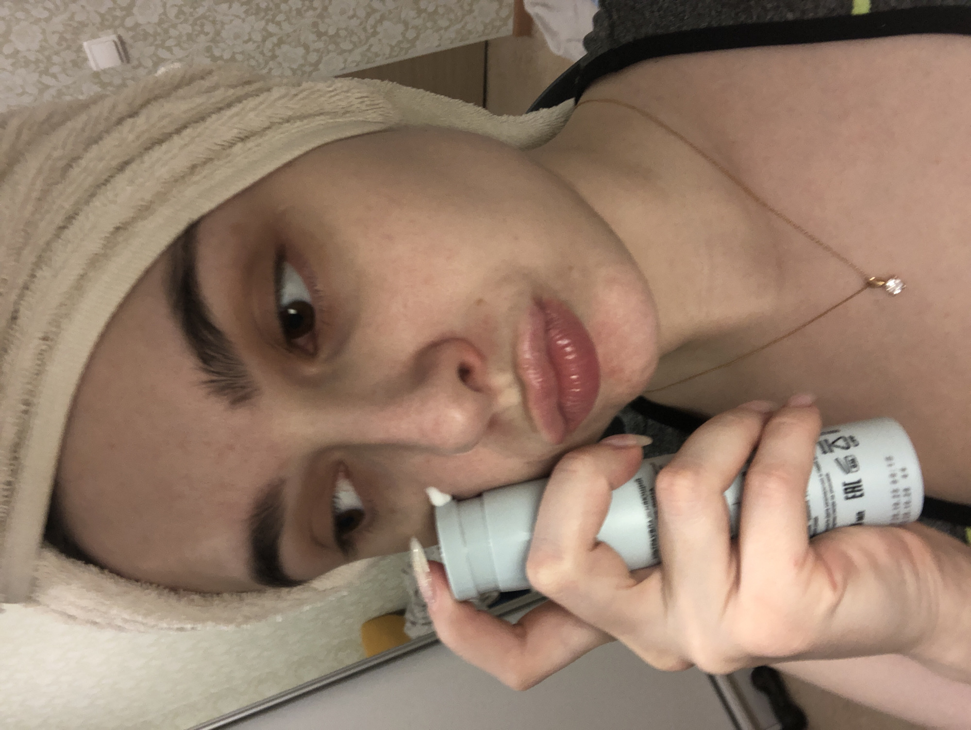 A woman with light skin and dark eyebrows is looking at the camera while holding a tube of cream near her face. She has a towel wrapped around her head and is wearing a necklace. The location is not discernible from the image.