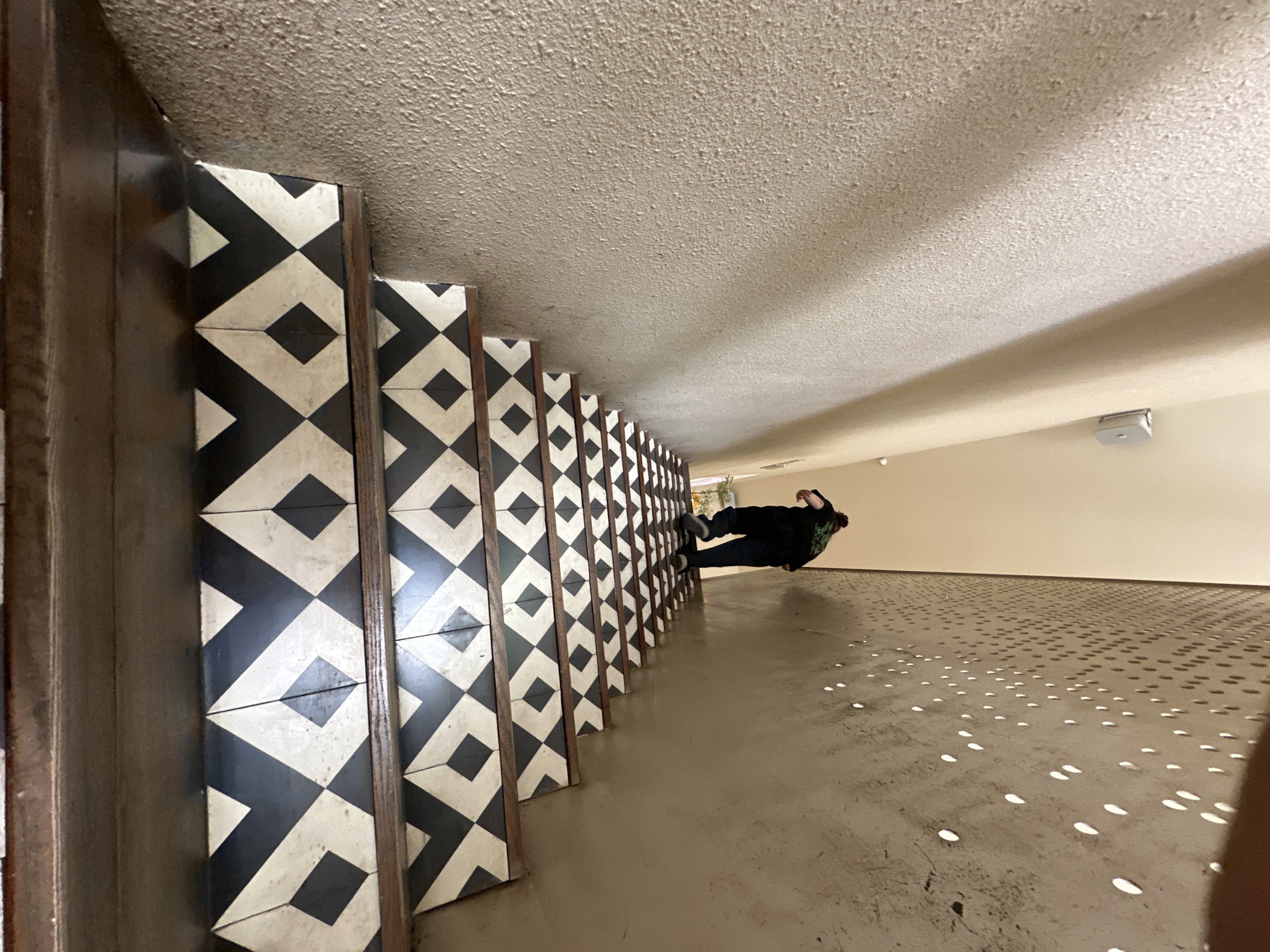 An individual is ascending a staircase. The staircase features a geometric pattern of black and white tiles on the risers and dark wood on the treads. The walls of the staircase are textured and appear to be painted a light, neutral color. On the right side of the staircase, there is a perforated metal wall, casting dappled shadows on the stairs and wall. The individual is wearing dark clothing and appears to be walking up the stairs. The location is in the city of Đà Nẵng, Vietnam.