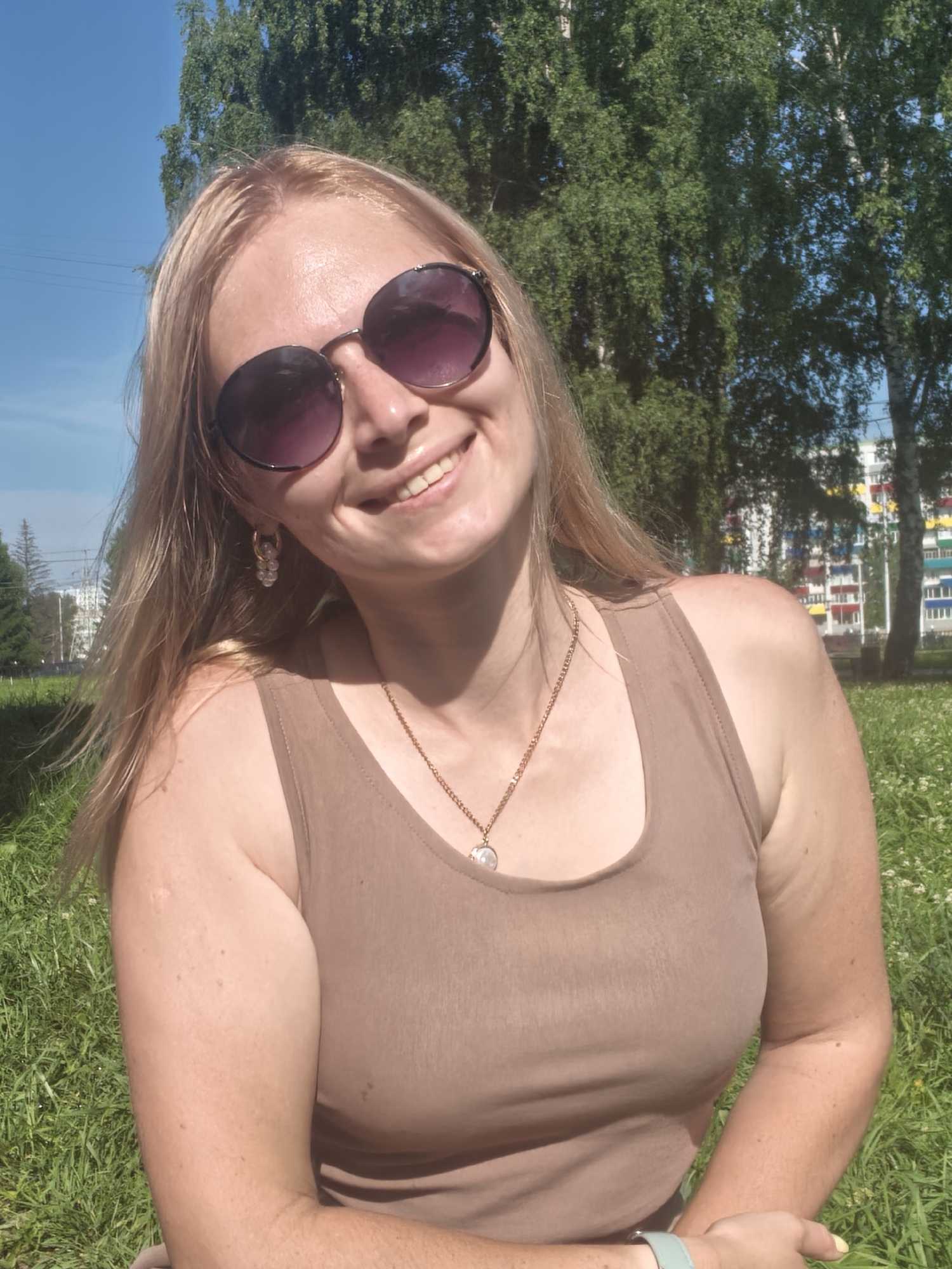 A medium shot captures a young woman from the chest up, outdoors on a sunny day. She has blonde hair, is wearing large, round sunglasses with purple lenses, and a light brown tank top. A gold chain with a pendant rests on her chest. She is smiling directly at the camera, with her head tilted slightly to the left. The background features green trees, a clear blue sky, and a building with colorful balconies, consistent with a residential area in Sterlitamak, Russia. The woman is seated on a grassy area with small white flowers.
