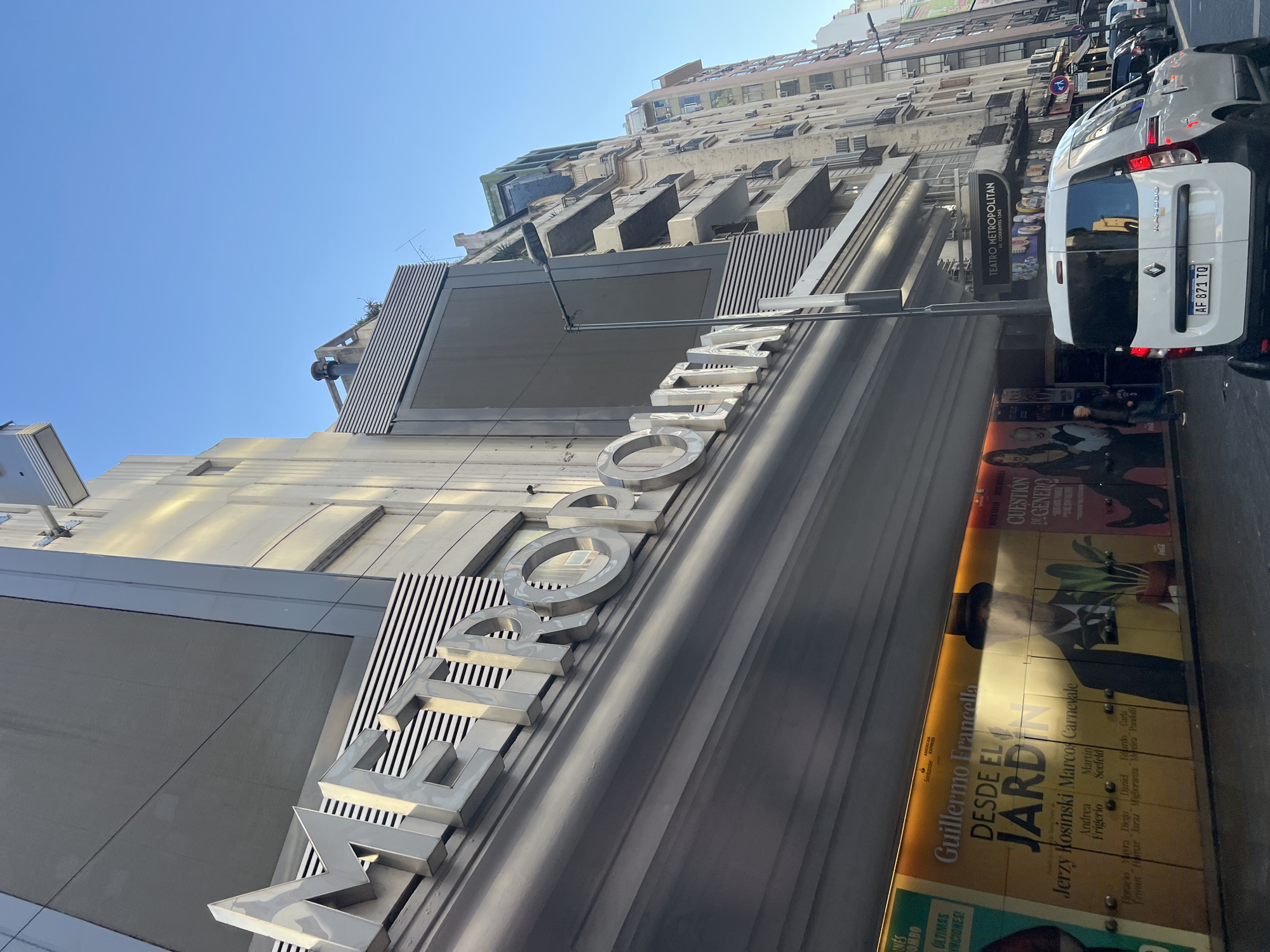 The image shows the Teatro Metropolitan Sura, located on Av. Corrientes 1343, in Buenos Aires, Argentina. The theater's name, "METROPOLITAN," is displayed in large, silver lettering on the building's facade. A series of movie posters are visible along the theater's entrance. A white Renault Kangoo van is parked beside the theater. The street scene also shows other cars and a sidewalk with pedestrians. Buildings are visible across the street. The sky is blue.