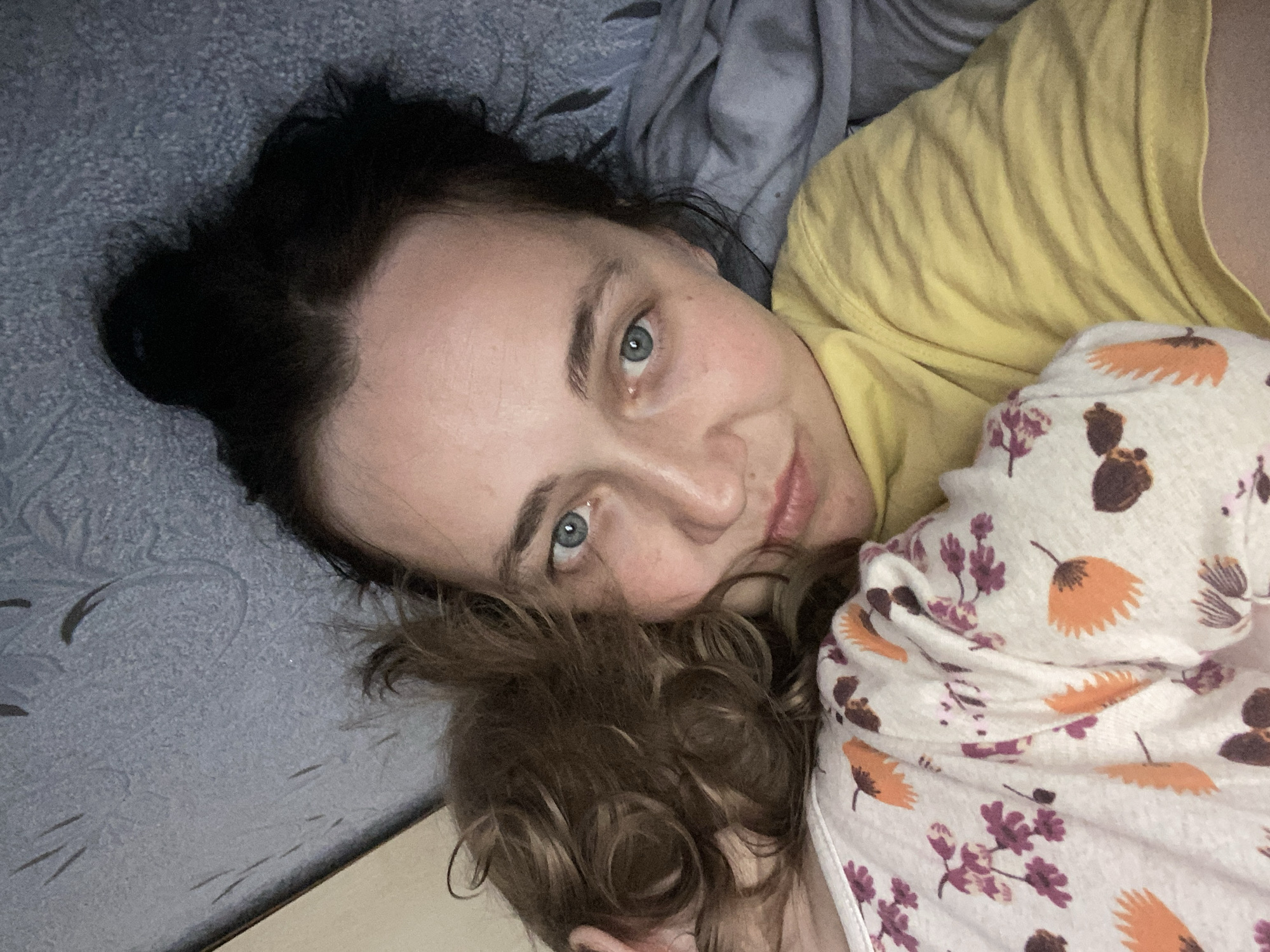 A young woman with dark hair pulled back and light blue eyes is lying down and hugging someone whose curly brown hair is visible. The woman is wearing a yellow t-shirt and the other person is wearing a white t-shirt with a floral and leaf pattern. They are in an indoor setting, possibly a bedroom, with a textured gray wall or headboard visible in the background and a gray blanket or sheet.