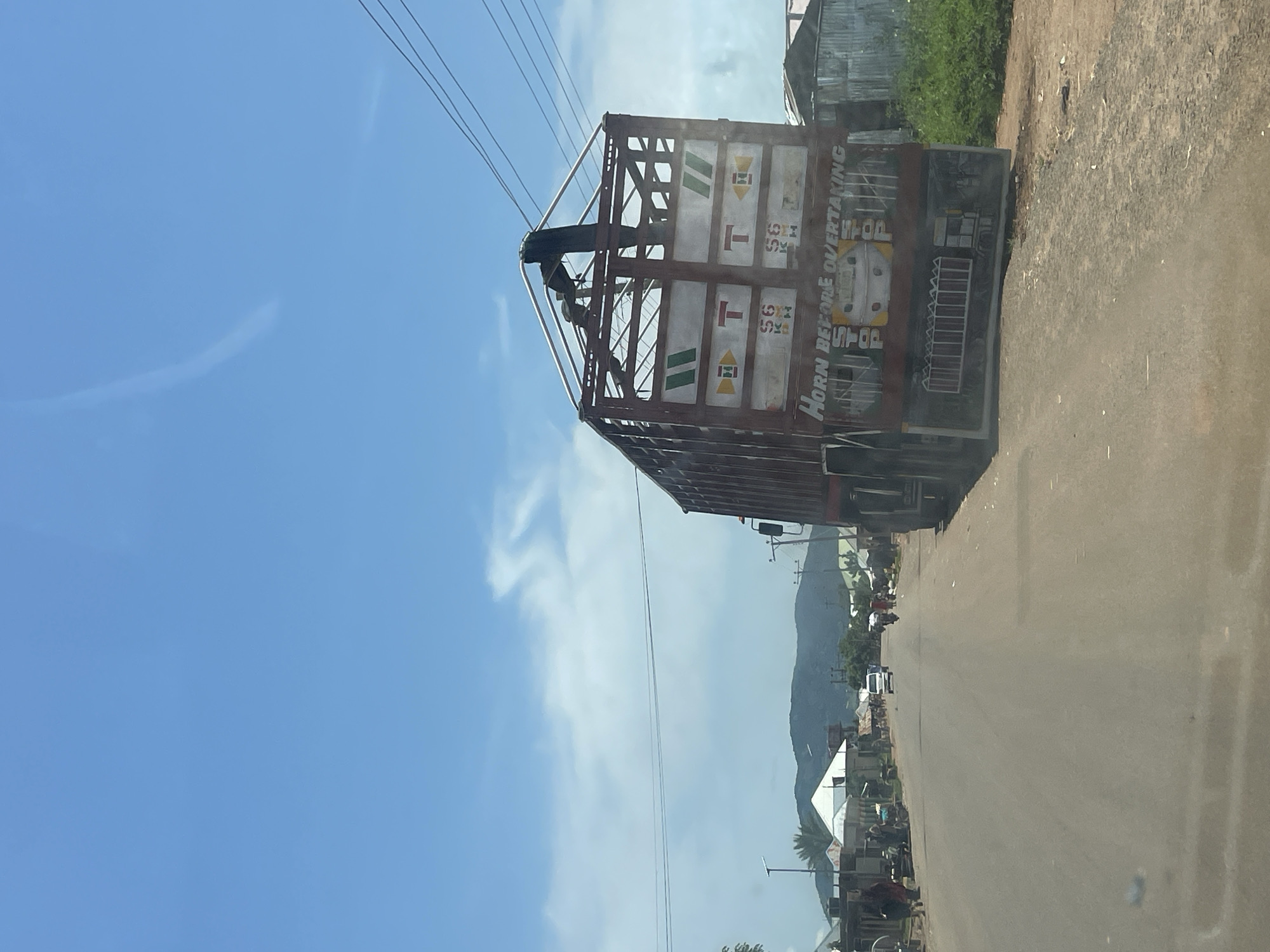 This is a photograph of a rural road in Zalau, Nigeria, under a bright blue sky with scattered clouds. A large, red truck is parked on the side of the road, its rear facing the camera. The truck has a white and yellow advertisement or logo on its side that says "HORN BEFORE OVERTAKING". The road is paved and appears to be a main thoroughfare for the community. On either side of the road are various buildings, including what appear to be shops and residences, some with corrugated metal roofs. There are a few people visible walking along the road and near the buildings, as well as some motorbikes and a white van. In the distance, rolling hills can be seen. The overall impression is one of a typical day in a bustling Nigerian town.