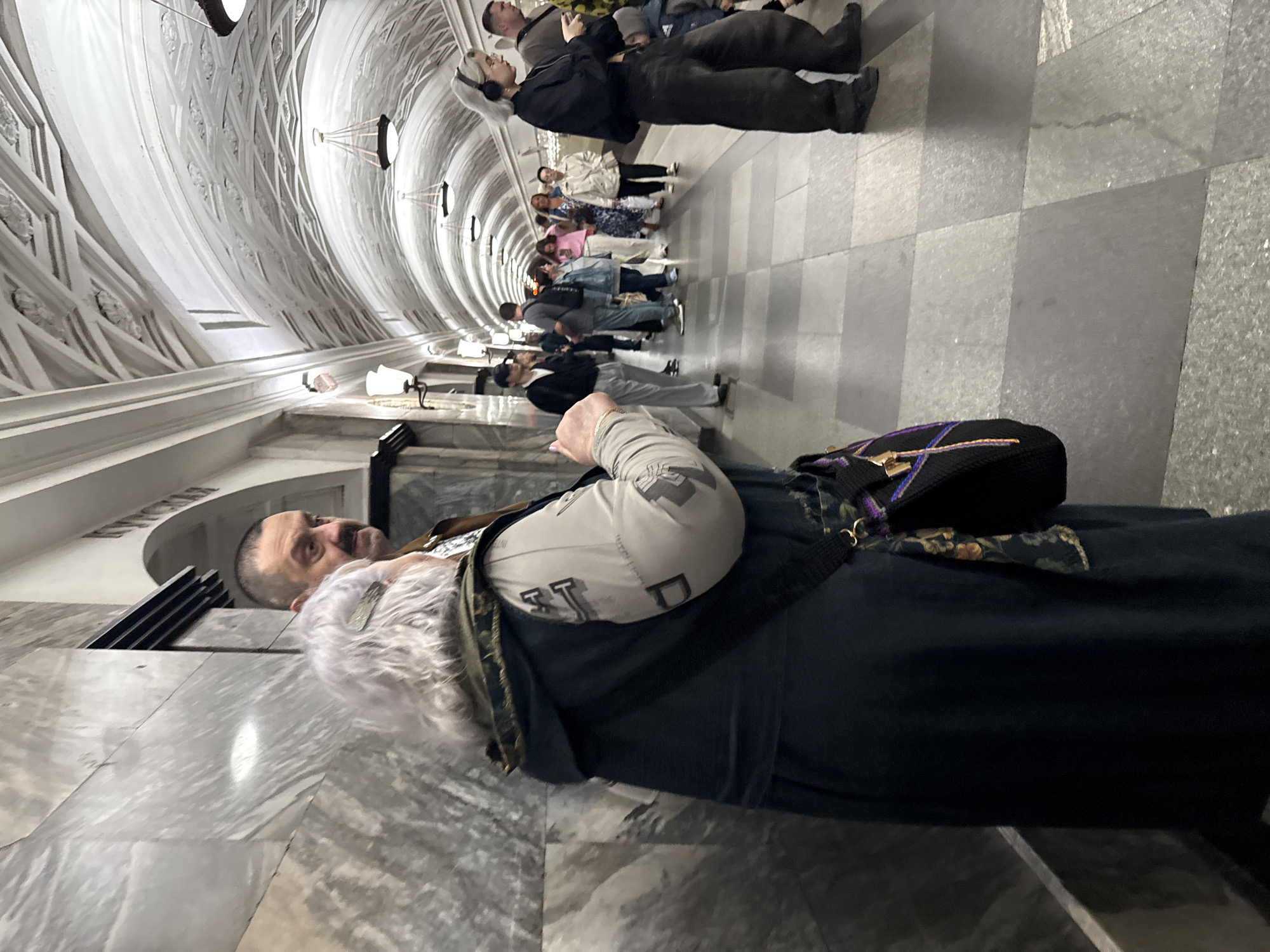 The image captures a view inside a Moscow Metro station, likely a platform or concourse, characterized by its distinctive vaulted ceiling adorned with ornate plasterwork and hanging chandeliers. Marble-clad walls line the side of the station. In the foreground, a woman with gray, permed hair and a dark hooded coat stands facing away from the camera, a patterned scarf peeking out from her bag. Beside her, a man with a mustache and short hair looks towards her. Further down the station, a diverse group of people are seen standing, some appearing to be waiting, while others are in motion. The floor is tiled with a checkered pattern of light and dark gray squares. The lighting is subdued, with the station's architectural features illuminated by strategically placed lamps. The overall scene suggests a typical day in the Moscow Metro, with commuters going about their routines.
