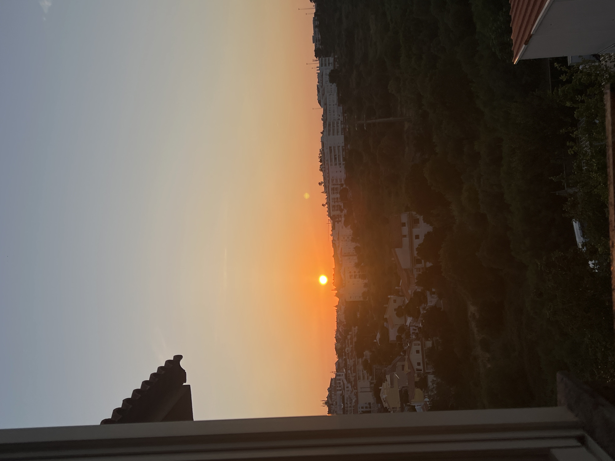 The image shows a sunset over a city that is part of Cascais, Portugal. The sun is low on the horizon, casting a warm orange glow across the sky. In the foreground, a building with a tiled roof is visible, with a window frame to the left. In the mid-ground, there are buildings with white facades, some of which appear to be apartment complexes. A hillside covered in dense green trees is behind the buildings. The sky transitions from a pale blue at the top to a soft orange near the horizon. There are no people visible in the image.