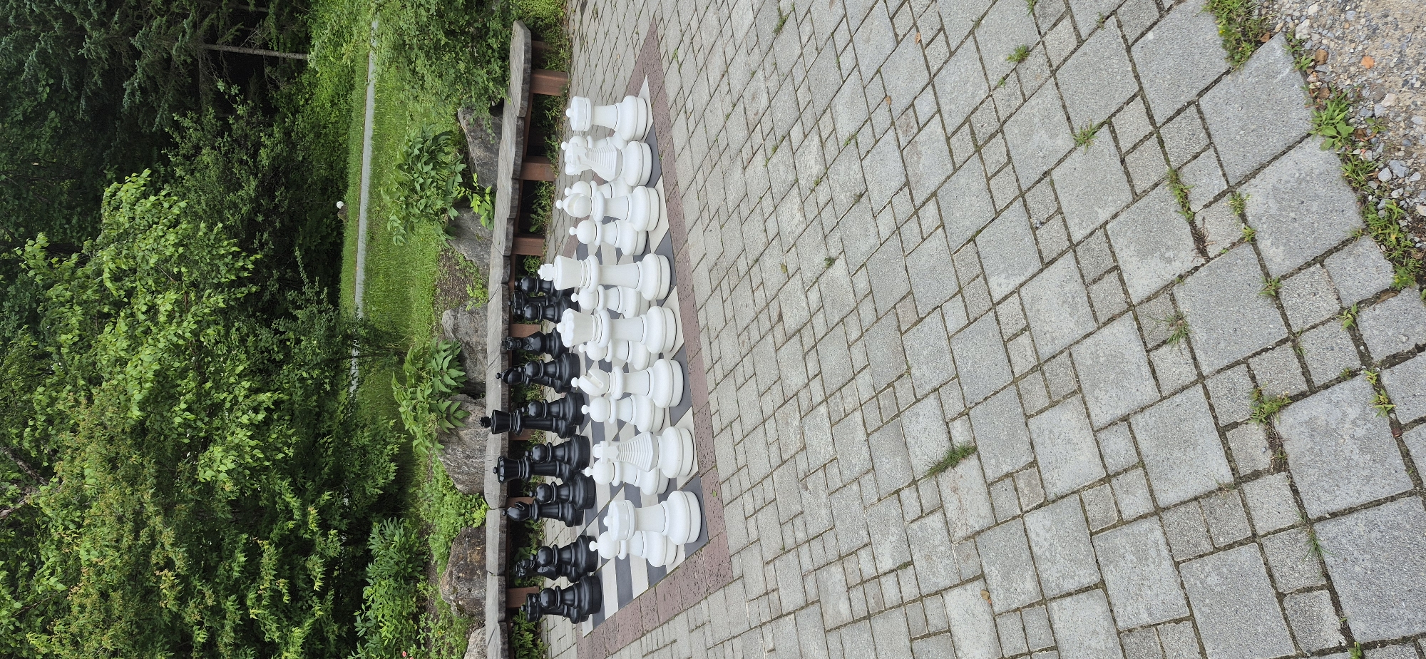 An outdoor giant chess set is positioned on a paved area in a park setting. The chess pieces are large, white, and black, and are arranged on the board in the starting position for a game. The chessboard itself is a grid of square tiles, some dark and some light, with a red border. Surrounding the chessboard is a paved walkway made of interlocking gray concrete pavers of varying sizes. In the background, there are wooden benches and natural landscaping, including lush green trees and bushes. The environment appears to be a recreational area within a city park, possibly Shtykovo, Russia, given the context. There are no people visible in the image.