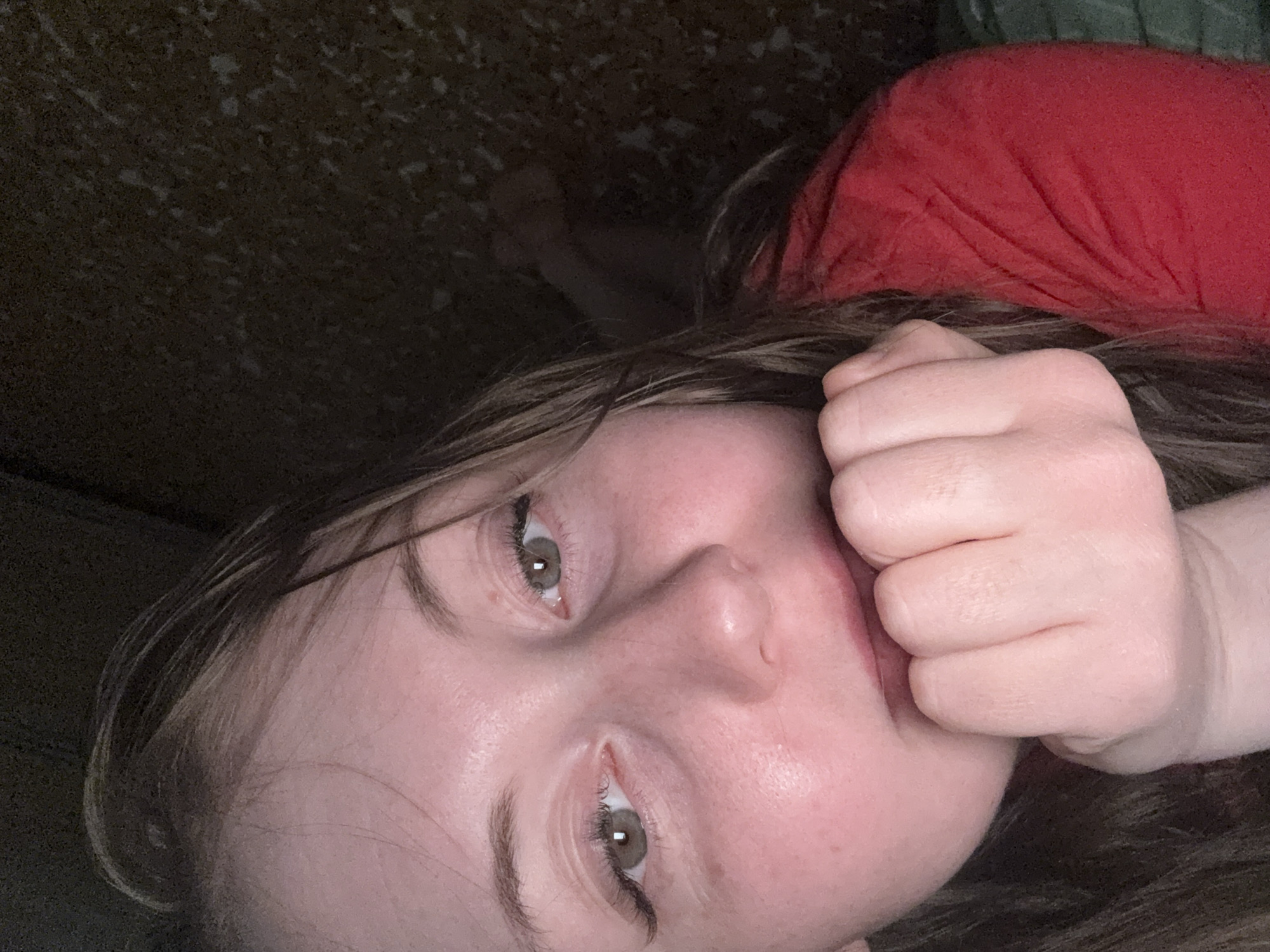 A young, fair-skinned person with brown hair and hazel eyes is lying down, appearing to be on a dark-colored couch or bed. They are wearing a red shirt and have their right hand partially covering their mouth. The location is Unknown, Unknown.