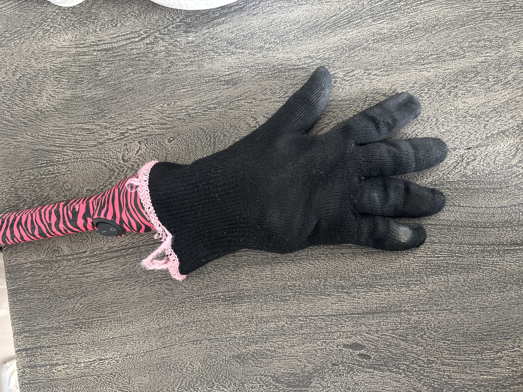 A black glove with a pink, zebra-striped handle and pink trim is lying on a textured, dark wood-grain surface. The glove is a hand-shaped covering, with distinct finger sections. The handle, attached to the wrist opening of the glove, features a pink and black zebra pattern and a black power button. The surface appears to be a tabletop. There are no people present, and no discernible signs of activity or interaction. The scene is static and appears to be indoors. The location is not specified in the image and cannot be determined.