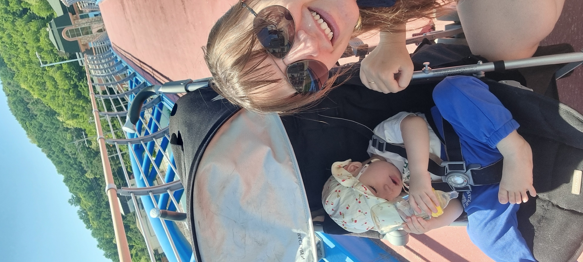 A woman and a baby are sitting in a stroller on a pink and blue structure on a sunny day. The woman is smiling and wearing sunglasses. The baby is wearing a hat and holding a bottle. The structure appears to be elevated with a complex design, with green trees and buildings visible in the background. The location is in Dunnae-myeon, South Korea.