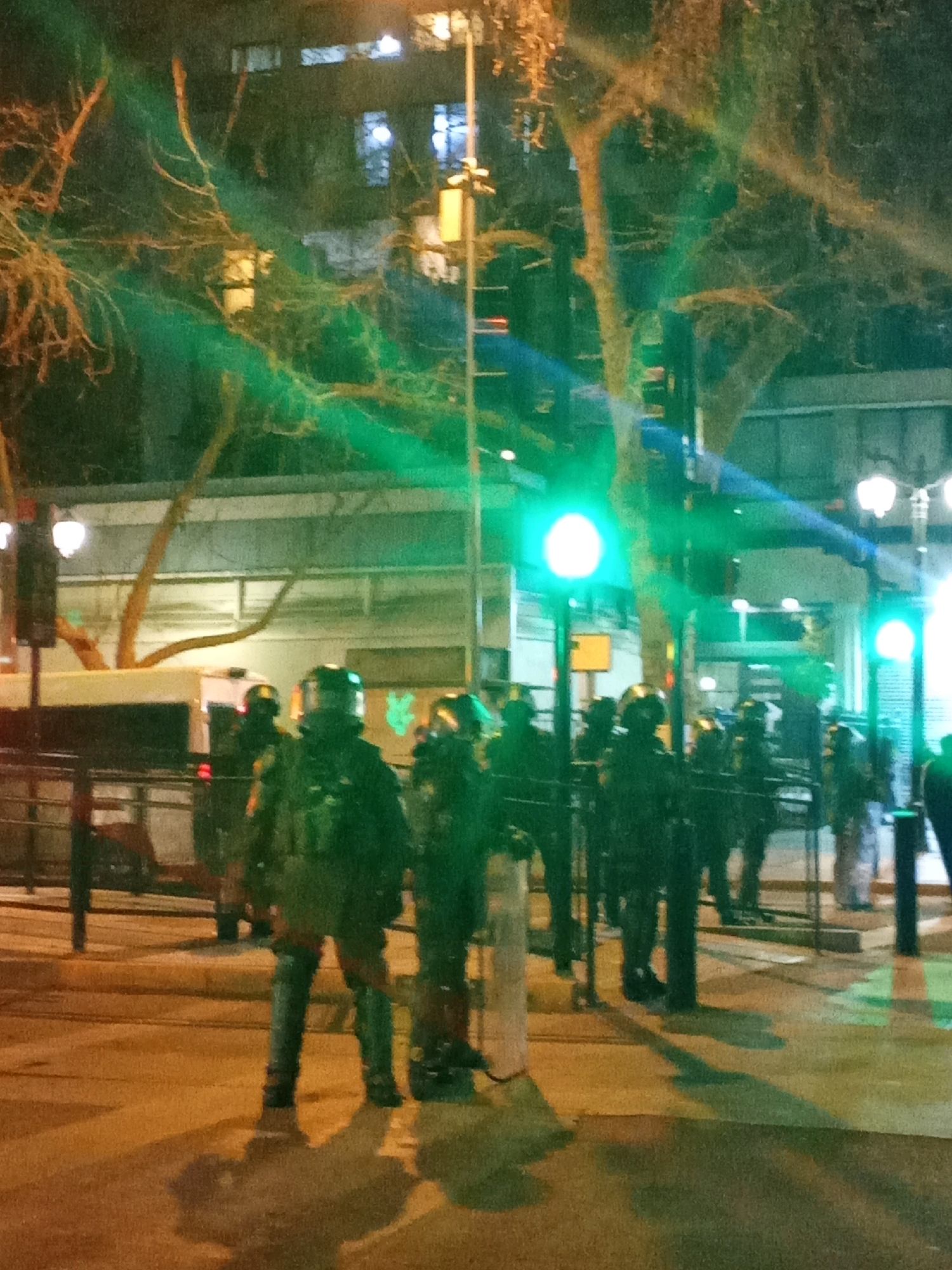 The image depicts a nighttime street scene in Providencia, Chile, with a group of individuals in riot gear, suggesting a potential protest or public order situation. The individuals are wearing dark uniforms, helmets with visors, and body armor. Some are holding shields, and they appear to be lined up, facing forward. There are traffic lights, streetlights, and trees visible in the background. The lighting is artificial, casting shadows and creating a somewhat hazy atmosphere, likely due to the low light conditions and possibly the presence of smoke or mist. A white vehicle, possibly a bus or police van, is partially visible on the left side of the frame. The ground is paved, and there are barriers or fences in the foreground. The overall impression is one of a controlled or tense environment.