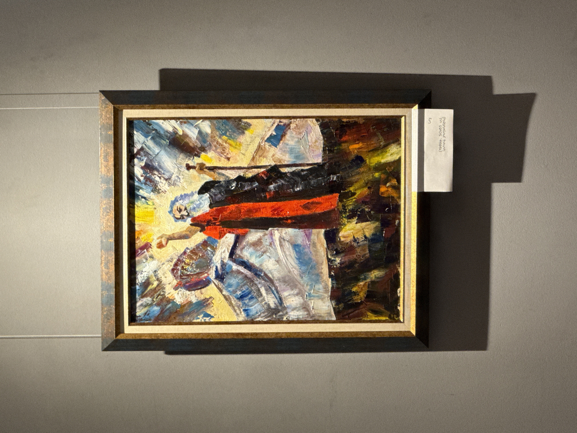 A medium shot depicts a framed painting displayed against a muted grey wall, possibly in an art gallery or museum. The painting, characterized by thick impasto brushstrokes in shades of yellow, blue, red, black, and white, features an abstract figure holding a long object. The frame is a double frame, with a dark outer frame and a gold inner frame. A small, white label is attached to the right side of the frame, with visible text in Armenian (upon translation it says "Narekian, Hrant (1950s uu before death)". Based on the name mentioned on the label, it is speculated that this painting is from the Hrant Narekian Art Gallery in Yerevan, Armenia. A horizontal wire is faintly visible on the wall to the left of the frame. The lighting creates a shadow behind the painting on the wall.