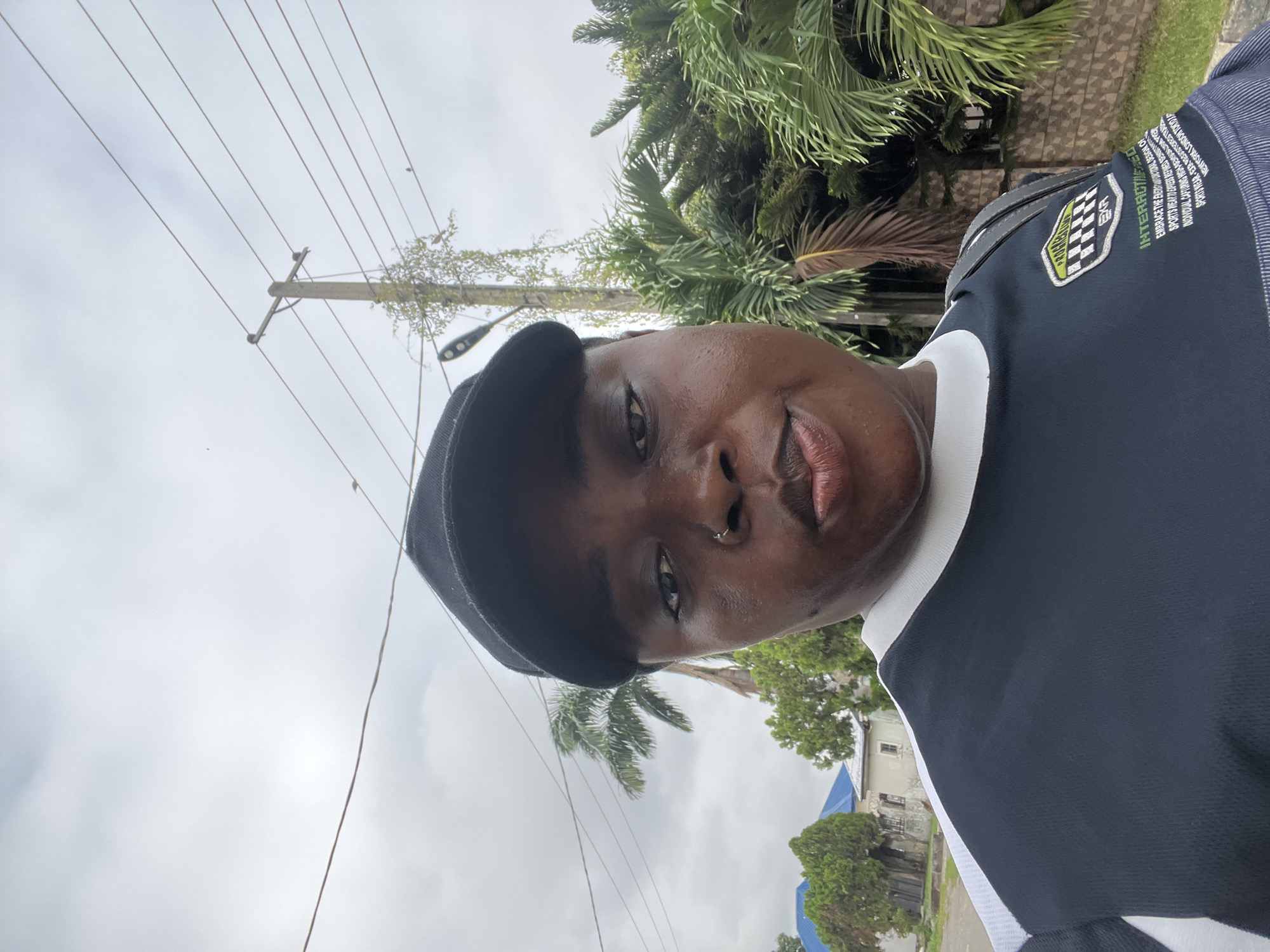 A medium-shot, outdoor selfie is captured in Abuja, Nigeria, under an overcast sky. A person wearing a black baseball cap and a dark blue shirt with white accents and a logo is facing the camera, looking directly at it. The person has dark skin, a nose ring, and their lips are slightly parted. Behind the subject, power lines stretch across the frame, supported by a wooden utility pole. Lush green palm trees and other foliage are visible, along with a glimpse of a building with blue roofing and a stone wall. The overall lighting suggests a cloudy day, and the scene appears to be an urban residential area.