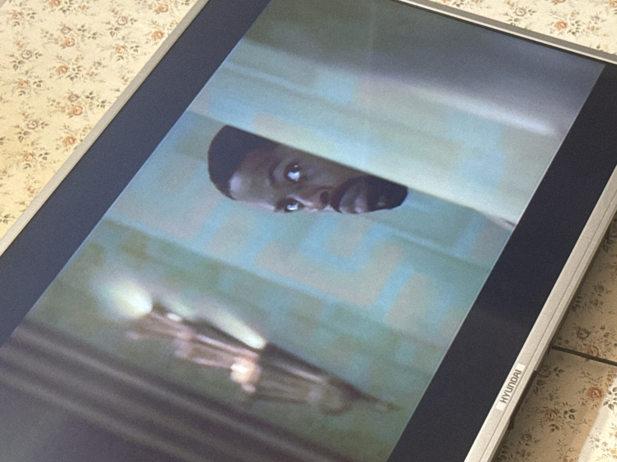 A photograph shows a Hyundai television screen displaying a scene of a Black man peering through what appears to be a slightly open door. The background behind the door is blurry, with hints of a chandelier-like object. The television is resting on a surface covered with a floral patterned tablecloth. The image was likely taken indoors. There is no information to suggest the location of Mytishchi, Russia.