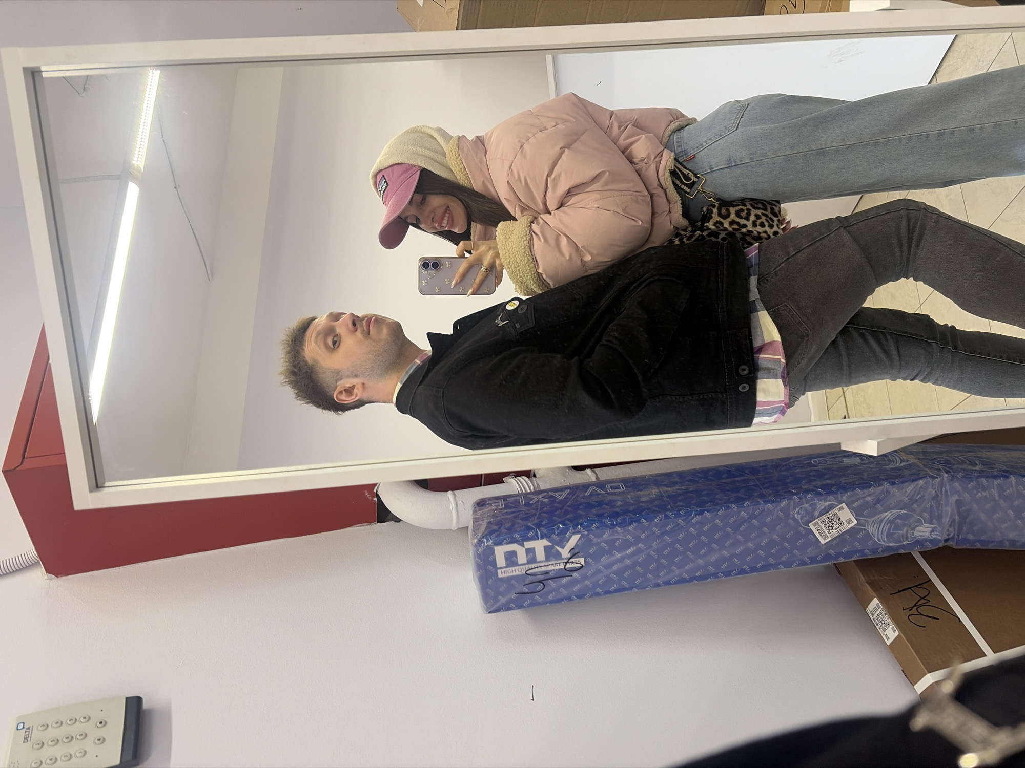 The image shows a selfie of a couple taken in a mirror. The man is wearing a black jacket, and the woman is wearing a pink puffer jacket and a pink baseball cap. The woman is holding a phone and taking the picture. In the background, there is a white wall, a red cabinet, a CV axle in packaging, and a security keypad. The location is likely in Saint Petersburg, Russia, given the Cyrillic alphabet on the CV axle packaging.