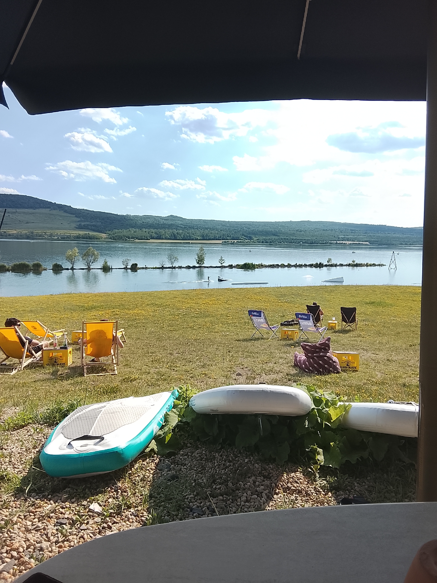 A serene lake scene unfolds under a bright, cloudy sky. In the foreground, a large, dark canopy partially obscures the view, framing the landscape. Two inflatable paddleboards rest on a bed of stones and greenery, one turquoise and white, the other white.
Beyond the paddleboards, a grassy area dotted with yellow flowers stretches towards the lake. Several people are scattered across this area, relaxing in brightly colored lawn chairs. Two yellow chairs are occupied, with individuals lounging, and two more chairs stand empty, one with a patterned pillow on it. Yellow crates are visible near the chairs, one with "CLUB-MATE" printed on it.
The lake itself is calm, reflecting the sky and the distant tree-lined hills. A series of small islands, adorned with trees, break the water's surface, creating a picturesque line across the lake. Further out, a wakeboard cable system with ramps and a tower suggests water sports are taking place, with a small boat visible in the distance. The time appears to be late afternoon, with the sun casting long shadows, and the weather is fair and warm. The overall atmosphere is one of peaceful recreation and leisure.
