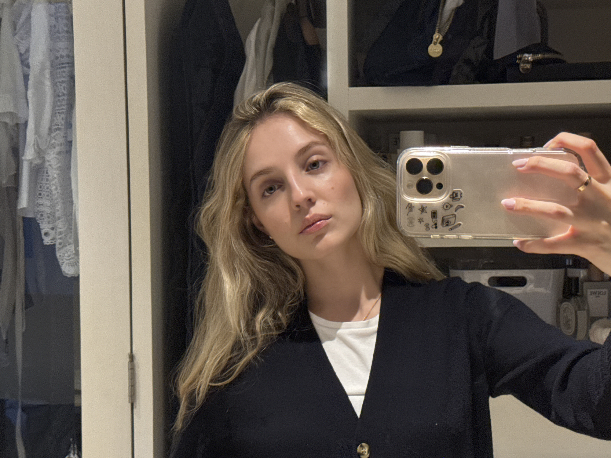 A blonde woman takes a selfie in a closet, holding a transparent phone case with stickers of drawings on it in her right hand. She is wearing a white t-shirt and a black cardigan. The closet is filled with clothes, and on the shelves above her, there are bags and bottles. The scene appears to be indoors, with no indication of location in Mexico City or any other city.