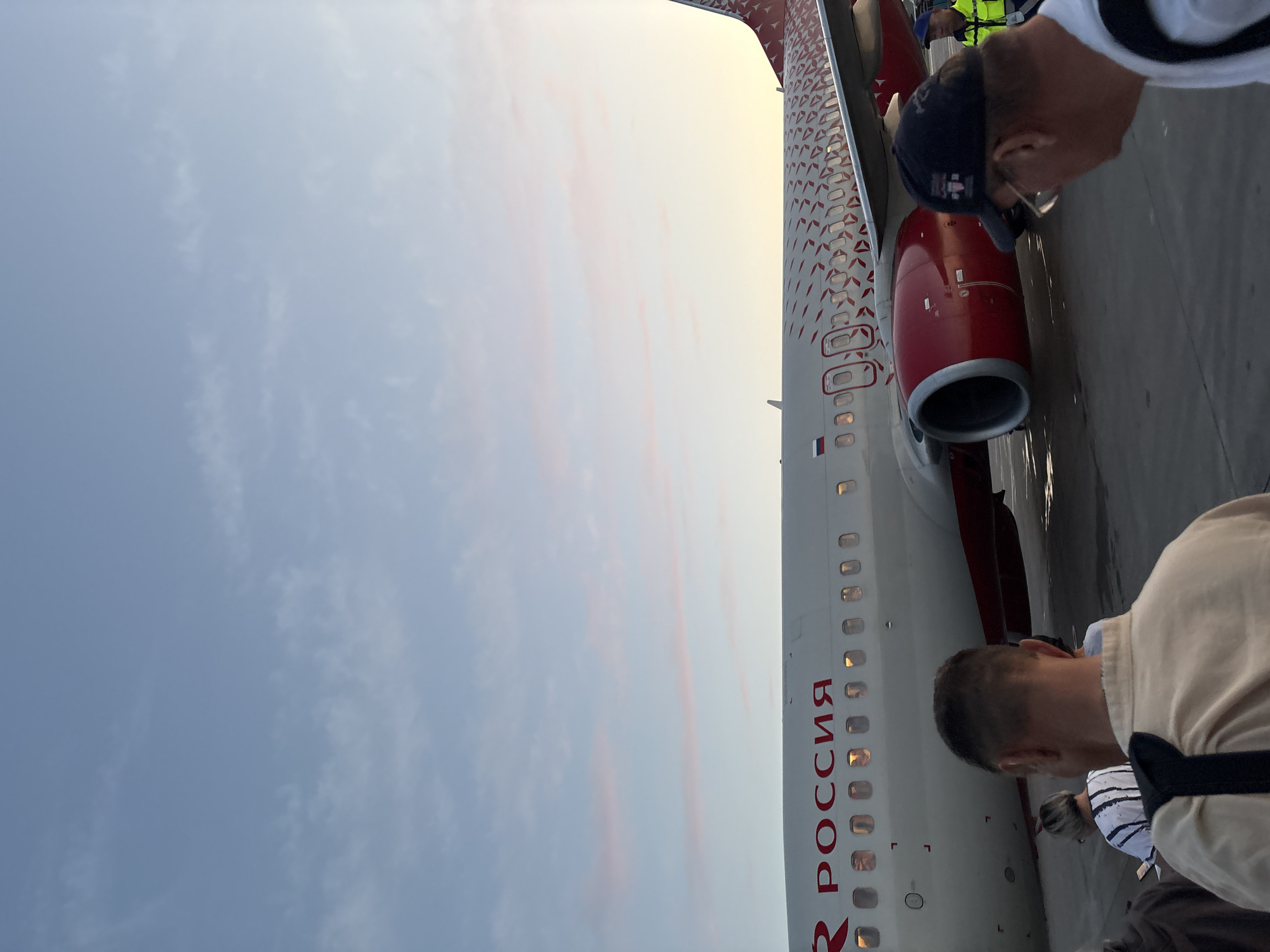 An airplane with the text "РОССИЯ" in red letters is parked on the tarmac at Bolshoye Savino Airport, Russia. The plane has a red and white patterned design on its tail and wing. Several people are visible on the tarmac, some walking towards the plane, suggesting boarding is in progress. The sky is light blue with wispy pink clouds, indicating either sunrise or sunset. The overall scene depicts an airport environment during a twilight period.