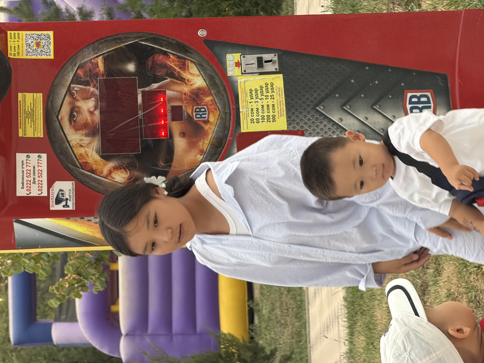 A medium shot captures two young children and a young girl standing in front of a red arcade game machine. The girl, positioned in the center, wears a white tank top under an unbuttoned light blue pinstripe shirt. She has dark hair pulled back and a white flower accessory in her hair, along with a white earring. To her right, a young boy in a white t-shirt with a black strap bag is slightly visible, looking down and to his left. In the foreground, a toddler wearing a white cap and a red shirt is partially visible on the far left, facing the boy in the white t-shirt. The arcade game, a punching game, features a graphic of a man's face and body with fiery accents. Red lights illuminate the score display. A yellow sticker with Cyrillic text and a QR code is affixed to the machine. The background shows a blurred purple and yellow inflatable structure, suggesting an outdoor amusement area with grass and trees. The overall scene appears to be set in a park or entertainment venue.