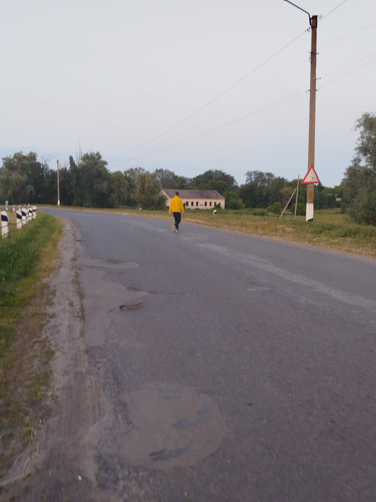 The image captures a rural paved road in Ustyvytsia, Ukraine, under an overcast sky. The asphalt road surface exhibits significant degradation, characterized by numerous visible potholes and cracks. On the left side of the road, a dirt shoulder transitions into a grassy verge, where a series of black and white striped roadside markers delineate the curve of the road as it recedes into the distance. Behind the vegetation on the left, a light-colored, single-story building with dark rectangular window openings is partially visible.
A lone individual, wearing a bright yellow long-sleeved top and dark trousers, is observed walking away from the camera, positioned on the center-right of the road. On the right side of the road, a utility pole, constructed from a light brown material, stands upright, supporting overhead power lines. Attached to this pole, approximately halfway up, is a red and white triangular warning sign, which appears to indicate "Uneven Road" (sign 1.16 in Ukrainian road signage). Dense vegetation, primarily consisting of trees and bushes, lines both sides of the roadway, forming a natural backdrop to the scene.