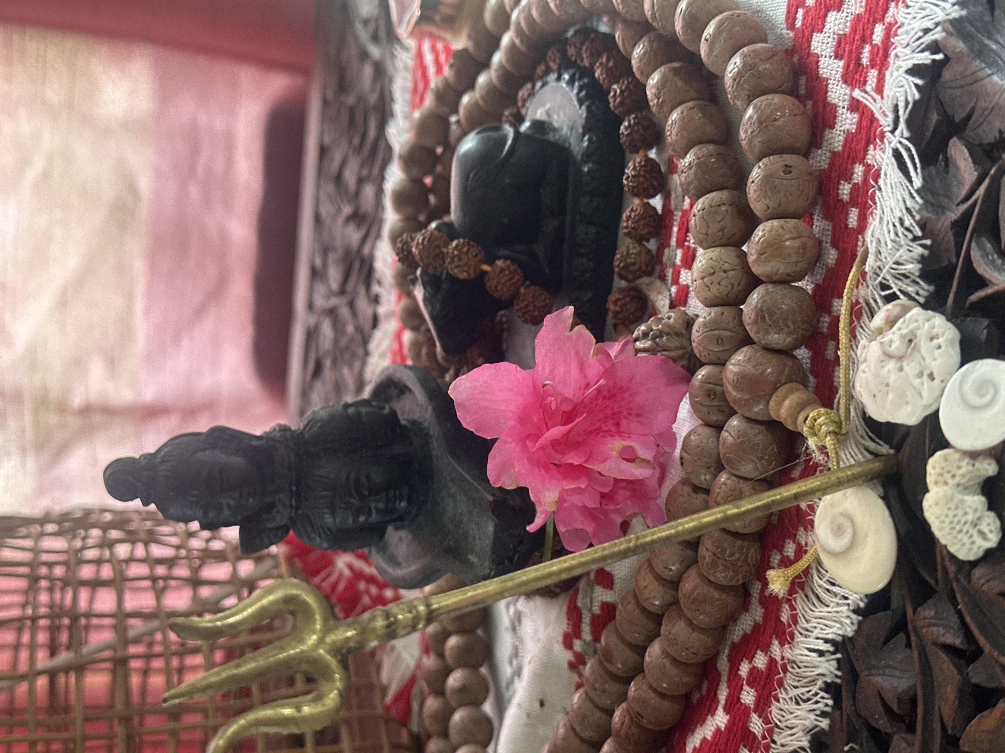 The image depicts a close-up view of religious items arranged on a surface. In the center, there is a black statue of a deity with multiple faces, possibly Lord Shiva. To the left of the statue, a golden trident, a symbol associated with Shiva, is prominently placed. Several strands of wooden prayer beads, likely mala beads, encircle the deity statue and extend outwards. A bright pink flower is positioned in front of the deity. Below the prayer beads and statue, there is a piece of white fabric with red embroidery and fringe. In the foreground, there are several small white objects, including what appear to be shells and coral fragments, placed on a dark wooden surface. The background is blurred, showing a woven basket-like structure and draped fabric in shades of pink and red. There is no information to suggest this scene is located in Thora, Australia.