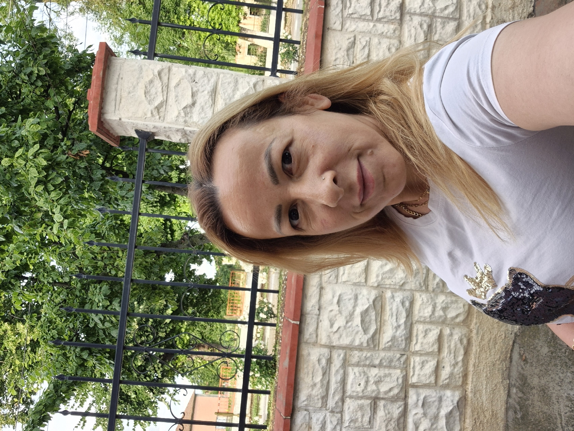 A woman with blonde hair stands outdoors, posing for a selfie. She is wearing a white t-shirt adorned with sequins in the shape of a deer's head. The background features a stone wall, a black metal fence with ornate details, and lush green trees. A stone pillar with a red roof separates sections of the fence.