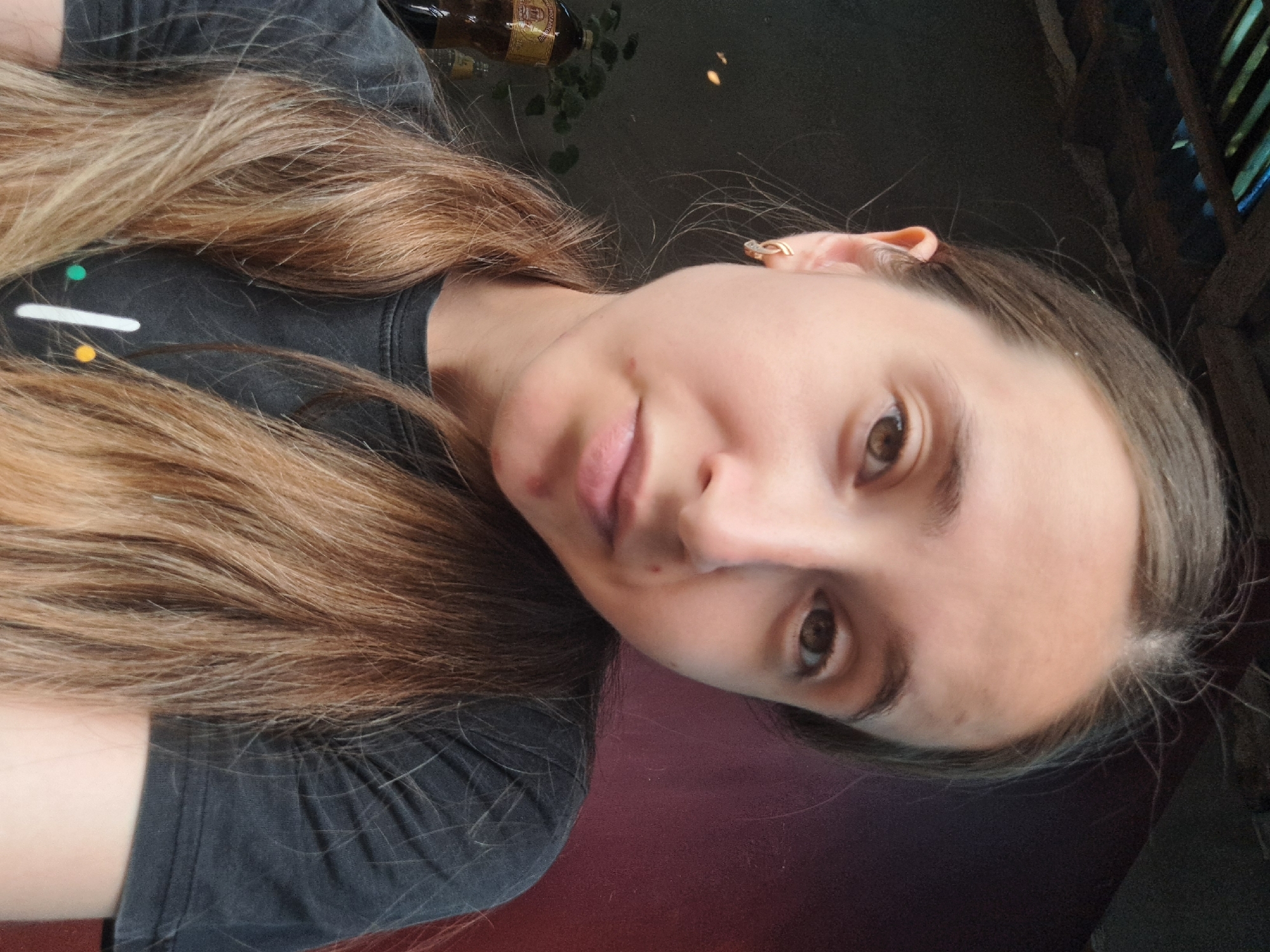 A young Caucasian woman with light brown hair tied in pigtails is looking directly at the camera. She is wearing a dark gray t-shirt with a white line and colorful dots on it. The background is dark, with a hint of red on the right side. A bottle and some greenery are visible on the left side of the image. The lighting suggests an indoor setting, possibly a home. The image quality is slightly grainy.