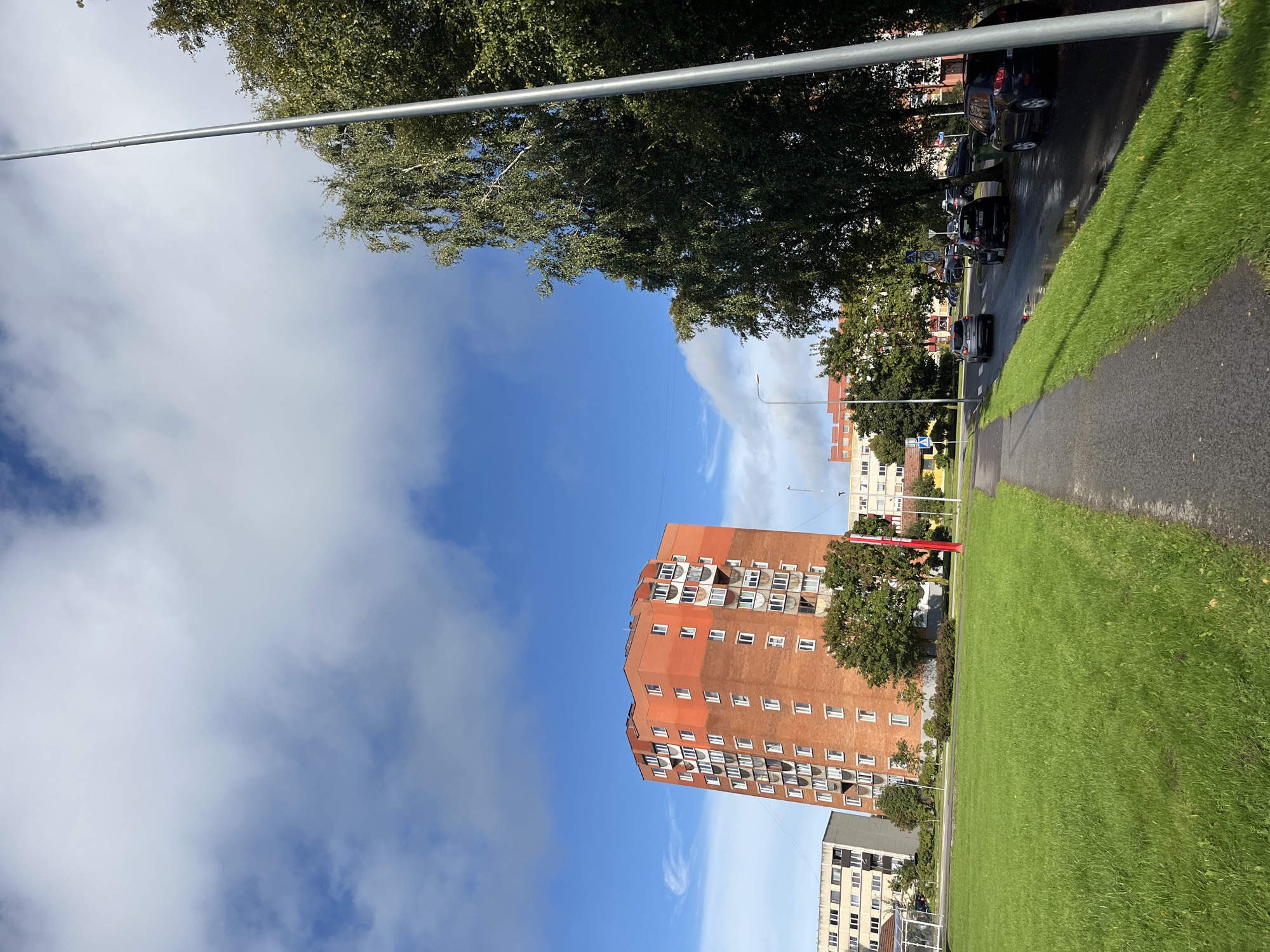 The image depicts a street scene in the city of Kohtla-Järve, Estonia, on a partly cloudy day. A tall, orange apartment building with many windows and balconies dominates the left side of the frame. To its left, a lower, light-colored apartment building is visible. A wide, green lawn with neatly trimmed grass occupies the foreground on the left. A paved sidewalk runs parallel to a wet asphalt road on the right, where several cars are parked and driving. Mature trees with green foliage line the right side of the road, partially obscuring other buildings in the background. A tall, silver lamppost stands prominently on the right edge of the image. The sky above is a mix of bright blue and white clouds.