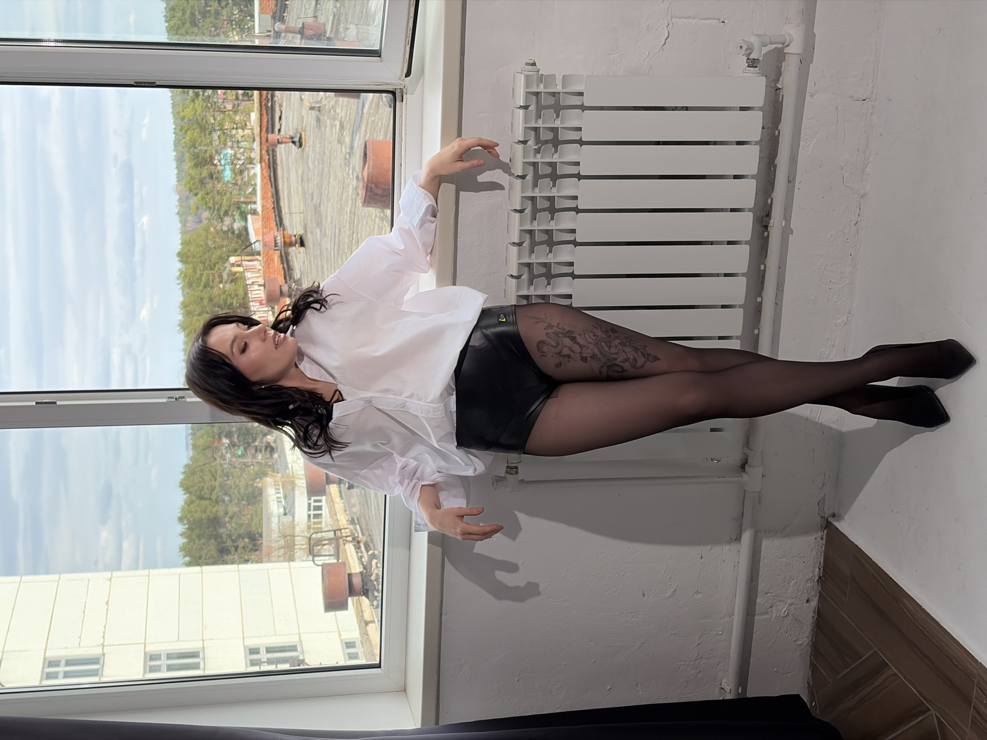 A woman with dark hair is posed sitting on a window sill near a radiator. She is wearing a white blouse, black shorts, black stockings with a floral pattern, and black high heels. The window looks out on a cityscape in Chaikovsky, Russia, showing buildings and trees under a cloudy sky. The walls are white, and there's a black curtain partially visible at the bottom of the frame.