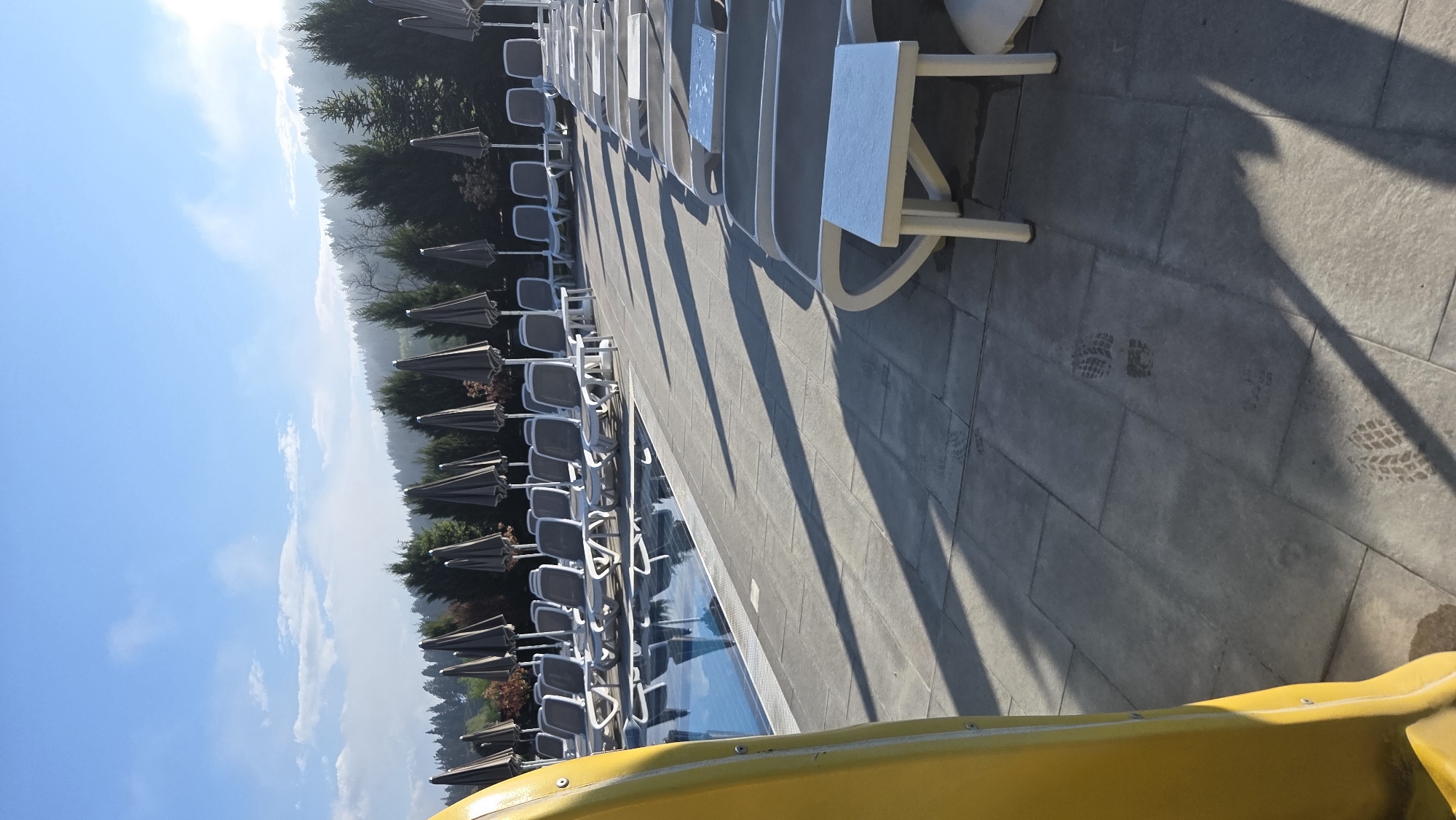 The image depicts a poolside scene with rows of lounge chairs and umbrellas. The foreground contains a section of a yellow water slide. A swimming pool is visible alongside the chairs. In the background, there's a line of trees extending towards a clear blue sky. Footprints are noticeable on the tiled patio area. The location is unspecified.