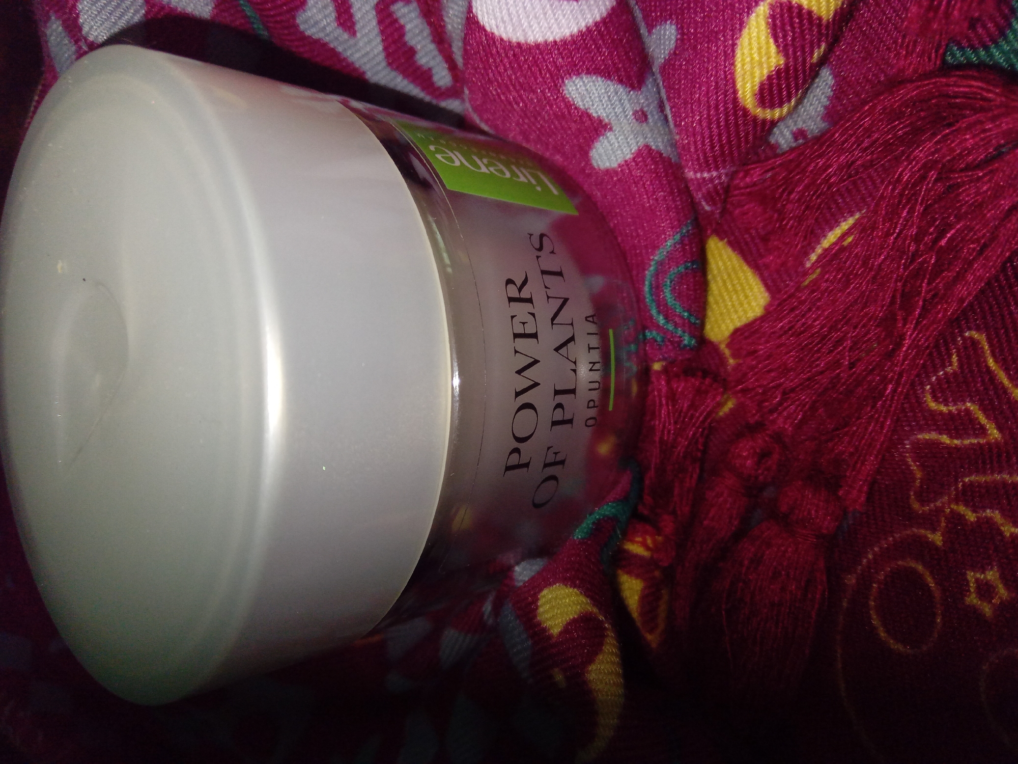 A close-up, overhead shot captures a jar of "POWER OF PLANTS OPUNTIA" cream, its lid a matte white and its container transparent, revealing the product inside. The jar is nestled within a vibrant fuchsia-colored fabric with a repeating floral pattern in white, yellow, and teal. To the right of the jar, a bright green sticker with the word "lirene" printed in white is visible. The fabric also features a fringe of deep red threads along the bottom edge of the frame. The lighting is dim, casting shadows and highlighting the textures of the fabric and the jar. The image is an indoor product shot with no indication of a specific location within Kitskany, Moldova.