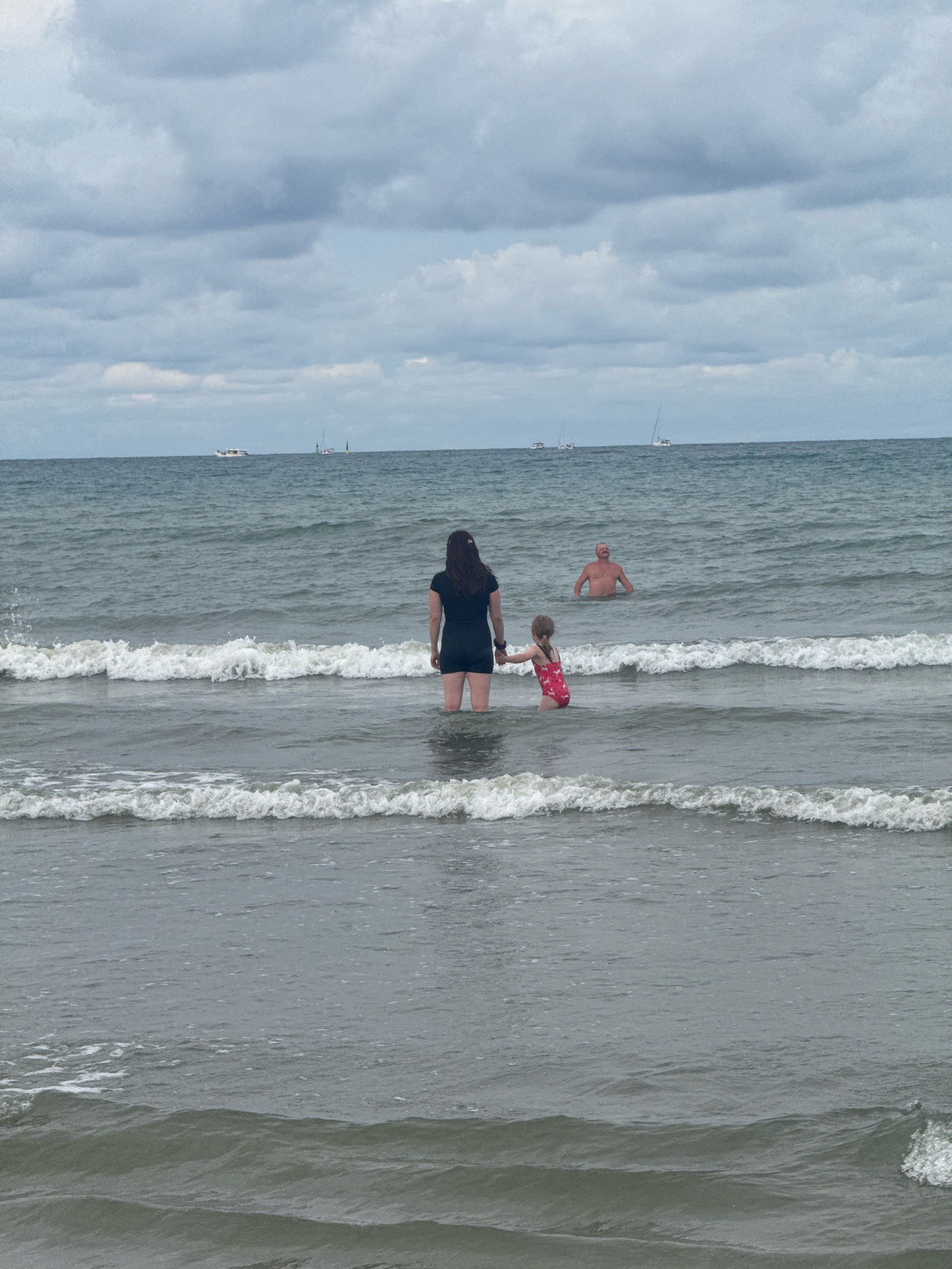 This media file depicts a beach scene with three people in the ocean. In the foreground, a woman with dark hair is standing in the water, wearing a black dress. She is holding hands with a young girl who is wearing a pink swimsuit with a floral pattern. Further out in the water, a man with a bald head is standing with his upper body visible. The ocean has waves breaking closer to the shore, and several sailboats are visible on the horizon under a cloudy sky. The lighting suggests it is daytime, and the cloudy conditions hint at overcast weather. The scene appears to be a family enjoying a day at the beach. There is no visible text in the image.