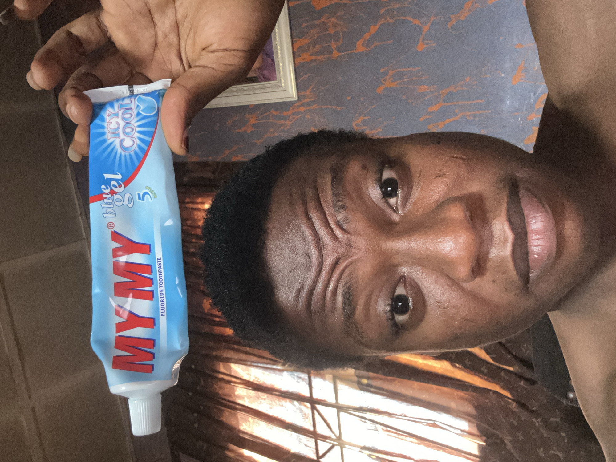 A medium shot of a dark-skinned person holding a tube of "MY MY" fluoride toothpaste. The person is positioned to the right of the toothpaste tube. The tube is light blue with red lettering and the words "Icy Cool" on the top. The background consists of a wall with orange splatter design and a brown curtain. The location is Unknown, Unknown.