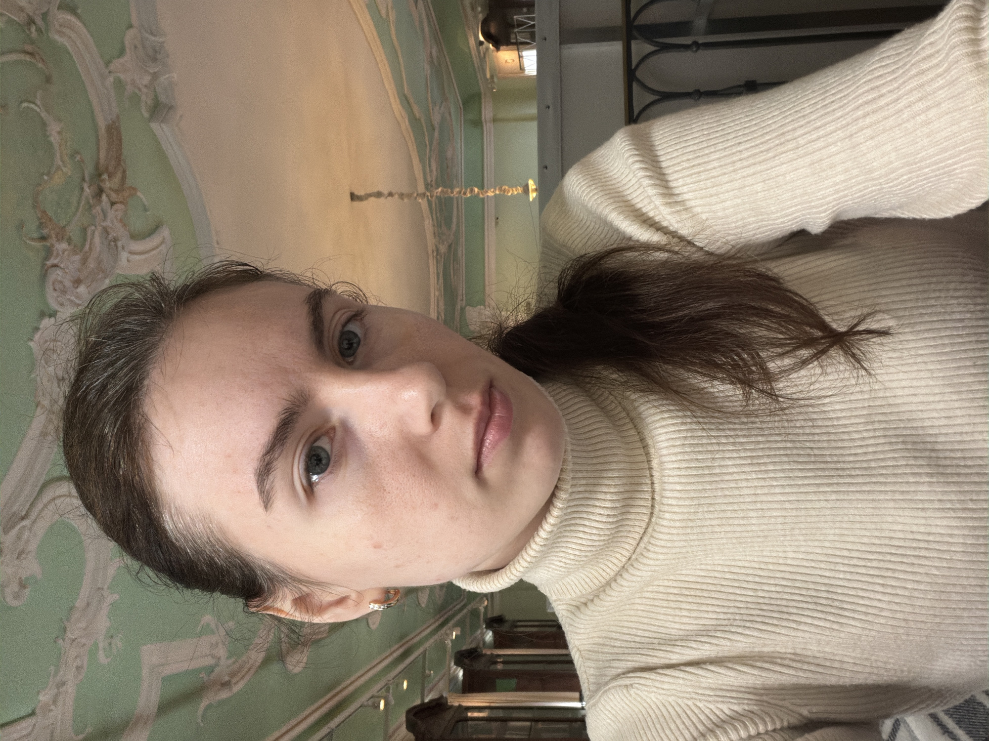 A young woman with light skin, blue eyes, and brown hair pulled back into a ponytail is posing for a selfie. She is wearing a cream-colored turtleneck sweater. The background shows ornate green and cream-colored wall decorations and part of a staircase. Based on the architectural style, the location is likely in Saint Petersburg, Russia.