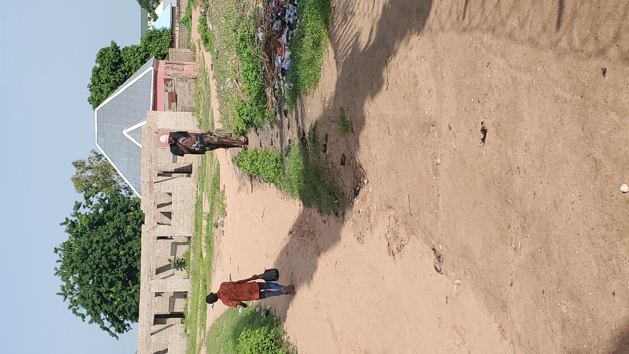 The image depicts a dirt road in Jalingo, Nigeria, with two individuals walking away from the camera. In the foreground, a young boy wearing an orange shirt and blue jeans carries a dark bag in his right hand. Further ahead on the right side of the road, an adult, possibly male, in a black t-shirt and shorts walks with a pink cap on their head, also carrying a bag. The environment is semi-arid, with sparse vegetation and some construction in the background. A partially constructed building made of concrete blocks is visible, along with a house with a corrugated metal roof. The road is unpaved, with visible tire tracks and scattered debris. The scene suggests an ordinary day in a developing urban or peri-urban area.