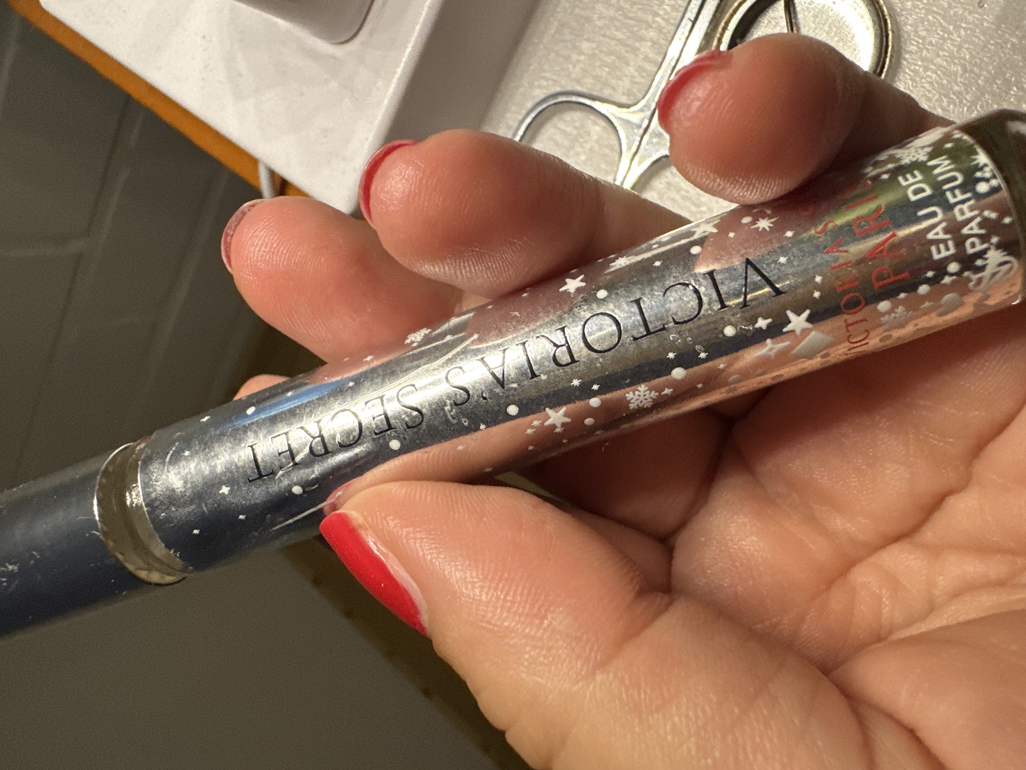 A close-up, eye-level shot shows a person's hand holding a small, metallic rollerball perfume bottle. The bottle is silver with a dark blue cap and is decorated with small white stars and snowflakes. Text on the bottle reads "VICTORIA'S SECRET PARIS EAU DE PARFUM." The person's fingers, painted with bright red nail polish, are visible supporting the bottle. In the blurred background, a pair of silver scissors and a white surface are discernible. The lighting suggests an indoor environment. The scene is devoid of any specific landmarks or context that would allow for its localization in Krefeld, Germany, or any other city.