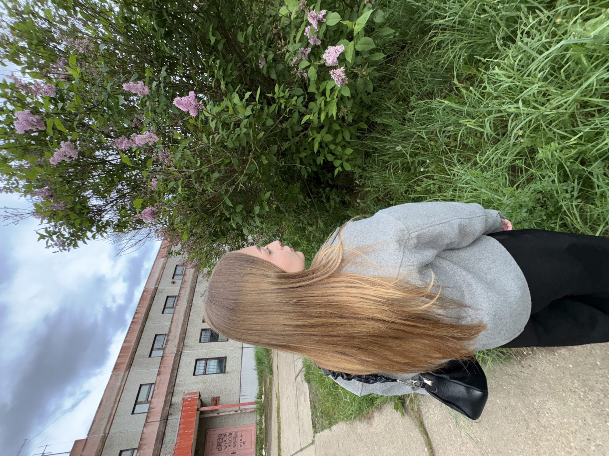 A young woman with long, light brown hair is shown in a photograph taken outdoors in what appears to be the city of Марковский, Russia. She is wearing a light gray sweatshirt and black pants. A black purse is visible next to her. She is positioned on a concrete surface next to grass. Behind her is a lilac bush with purple flowers. To the left, a tall, older building with multiple windows is partially visible. The sky is overcast.