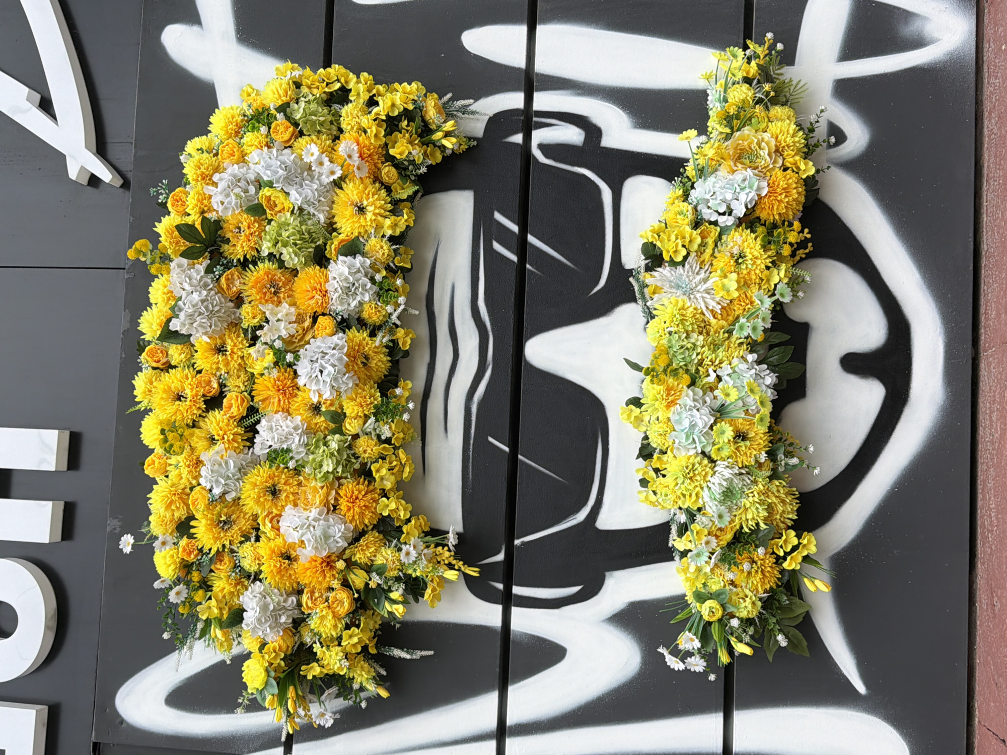 This media file displays a close-up, overhead view of two large, decorative floral arrangements. Both arrangements are primarily composed of yellow and white flowers, with some touches of green foliage and lighter green hydrangeas. The flowers vary in size and texture, with prominent yellow pom-pom-like blooms, smaller daisy-like flowers, and clusters of hydrangeas.
The floral arrangements are affixed to a dark grey wooden surface, which appears to be made of vertical planks. This surface is decorated with a large, stylized black and white graphic. The graphic has a bold, graffiti-like appearance with sweeping lines and rounded shapes, vaguely resembling abstract figures or perhaps oversized letters.
On the left side of the image, partially visible, are several three-dimensional white letters attached to the grey wooden surface. The letters appear to be a capital "Y" and parts of other letters, with a thick, blocky font.
The lighting suggests it is daytime, and the overall scene is static, with no discernible activity taking place beyond the display of the floral art. The focus is on the vibrant colors and textures of the flowers against the stark background. There is no visible text other than the white letters on the left.