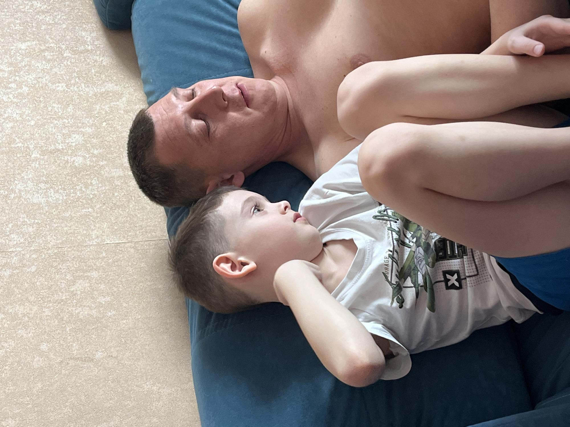 A shirtless man and a young boy are lying together on a blue couch. The man's eyes are closed, while the boy, wearing a white t-shirt and blue shorts, is looking upwards with his head resting on the man's arm. The scene takes place indoors, with a beige tiled floor visible to the left. There is no information in the provided image or prompt that specifies this location to be in Chaikovsky, Russia.
