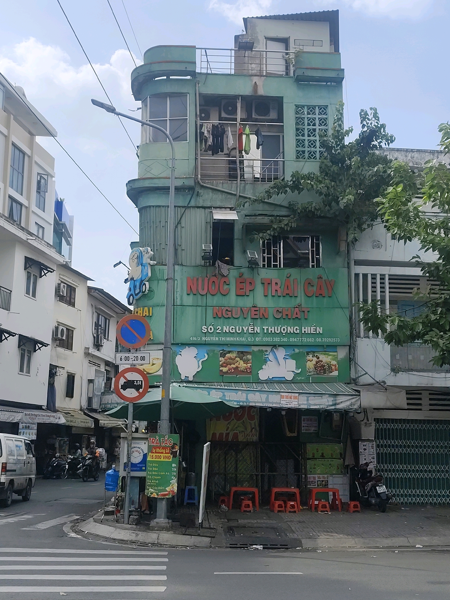 This is a street-level, eye-level, medium shot of a busy intersection in Ho Chi Minh City, Vietnam. The scene is dominated by a multi-story building with a green facade, featuring signs for a fruit juice shop. The building's architecture suggests a mix of older and newer construction, with balconies adorned with drying clothes and air conditioning units.
Several street signs are visible, including a "no parking" sign with a blue circle and red diagonal line, a sign indicating "6:00-20:00," and a "no trucks over 3.5 tons" sign. A lamppost with wires running across it is positioned near the corner.
On the street level, there are several motorbikes, a white van, and a blue water cooler. Red plastic stools are arranged outside the juice shop, indicating an area for customers. The pavement is cracked and shows signs of wear. The overall environment is typical of an urban Vietnamese street scene, with a blend of commercial activity and residential elements. The weather appears to be overcast.