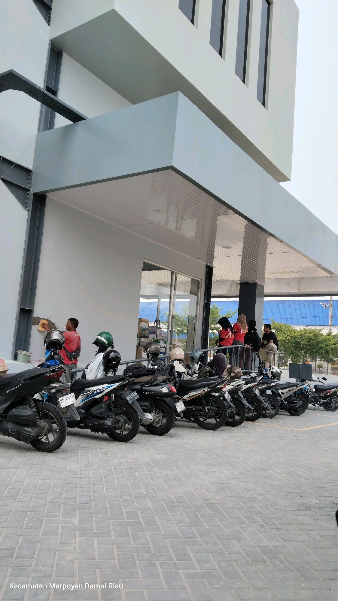 A wide shot captures the exterior of a modern building with a multi-level overhang, likely a commercial establishment, in Pekanbaru, Indonesia. The building's facade is primarily white and gray, featuring large, dark-paned windows and angular architectural elements. In the foreground, a row of parked motorcycles, predominantly scooters, lines a paved courtyard. The ground is paved with interlocking gray brick pavers arranged in a herringbone pattern.
Several individuals are present. On the left, a young man in a red shirt is actively applying plaster to the building's exterior wall with a trowel, indicating ongoing construction or maintenance. To his right, a few other individuals are standing or leaning against a railing, seemingly observing or waiting. Further to the right, a group of three women, two in red shirts and headscarves and one in a beige headscarf, are interacting near the railing. On the far right, a man in a dark shirt is also standing. Helmets are visible on some of the parked motorcycles. The background reveals a glimpse of a blue-roofed structure and some trees, suggesting an urban or semi-urban setting. The text "Kecamatan Marpoyan Damai Riau" at the bottom left corner of the image indicates the specific administrative region within Pekanbaru. The sky is overcast, suggesting a diffused light throughout the scene.
