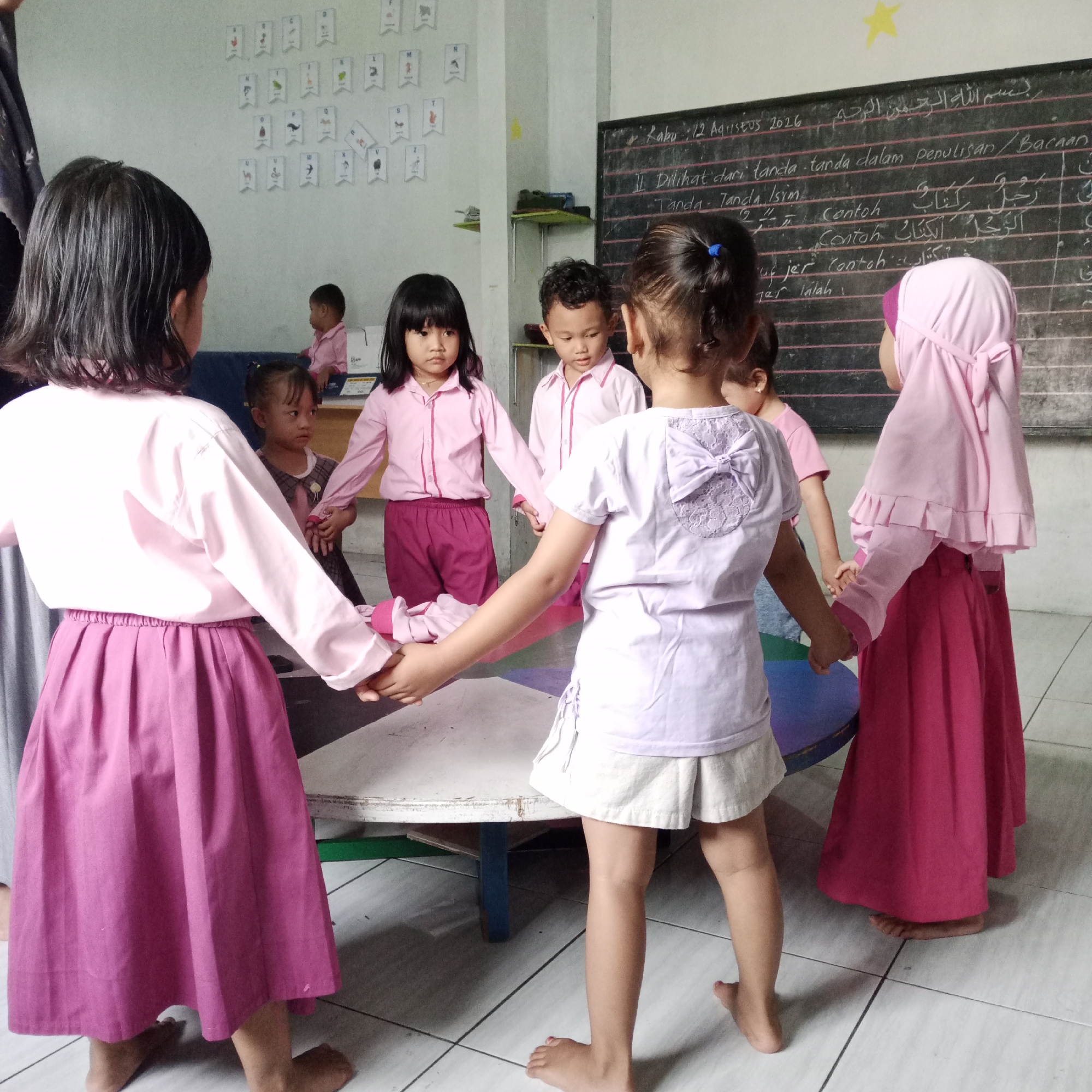 In a classroom in Rejosari, Indonesia, a group of five young children are holding hands in a circle around a low, round table. The children are dressed in pink and purple uniforms. A blackboard is visible in the background with some writing in Arabic. There are also flashcards with letters and images on the wall. The floor is tiled in a light color.