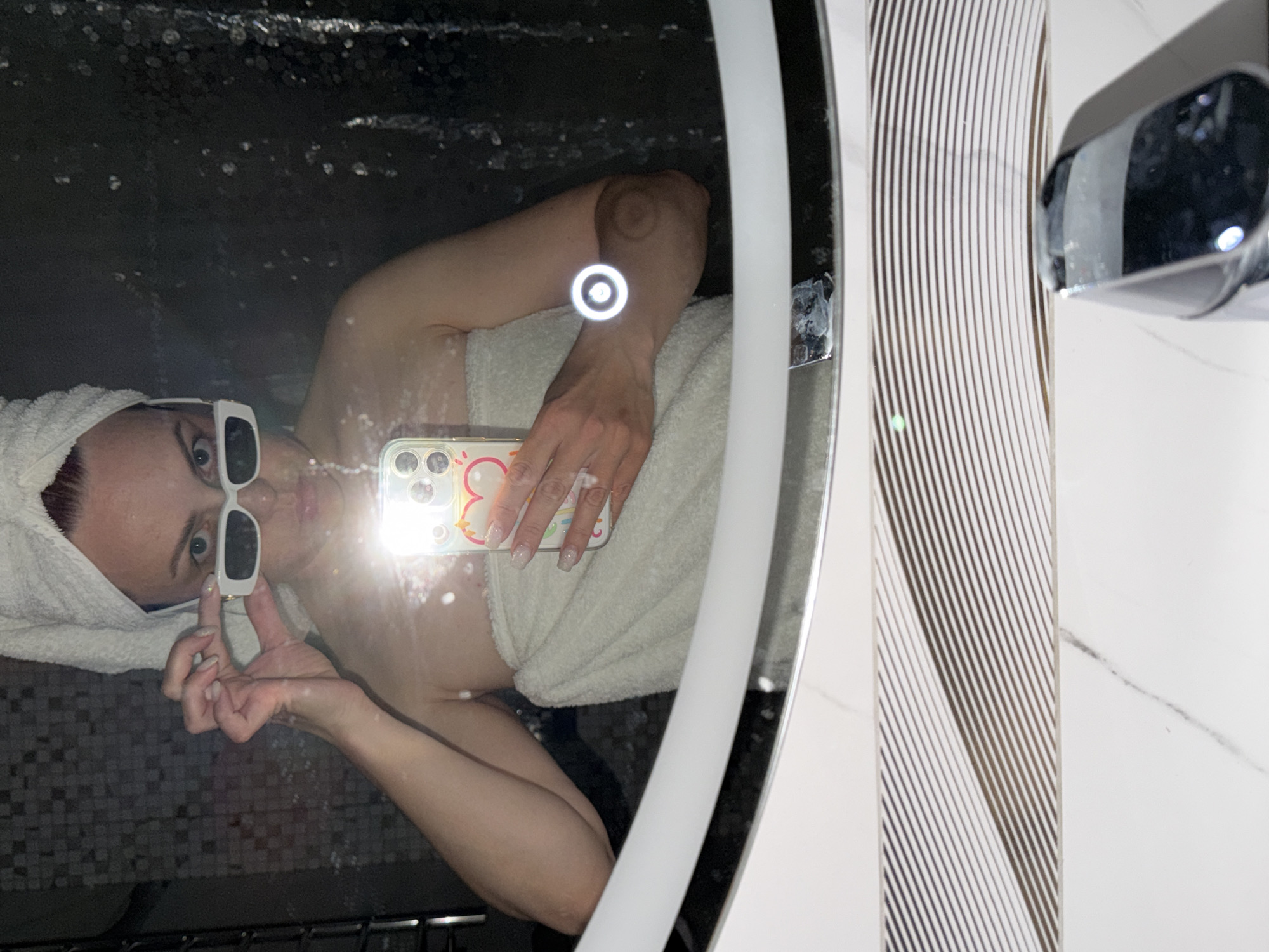 A woman is reflected in a bathroom mirror. She is wearing a white towel on her head and white sunglasses. She is holding a phone with a decorative case. The bathroom has white and grey striped tiles, and a chrome fixture is visible on the right. The location is a bathroom, possibly in Votkinsk, Russia, given the query.