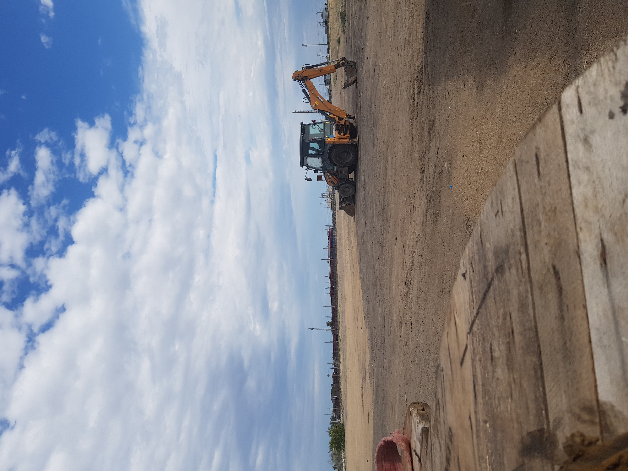 The image depicts an outdoor scene in the city of Kurchatov, Kazakhstan. In the foreground, a portion of a large, weathered wooden structure, possibly a spool for industrial cable, is visible. The middle ground is dominated by a dirt lot with a backhoe loader parked in the center. The backhoe is orange and black, with its front loader and backhoe arm both extended. In the background, a long train with red and blue cars is stationary on a track. Scattered utility poles and some sparse vegetation are also present. The sky is partly cloudy with a mix of blue sky and white clouds. The overall impression is one of an industrial or construction area under a partly cloudy sky.