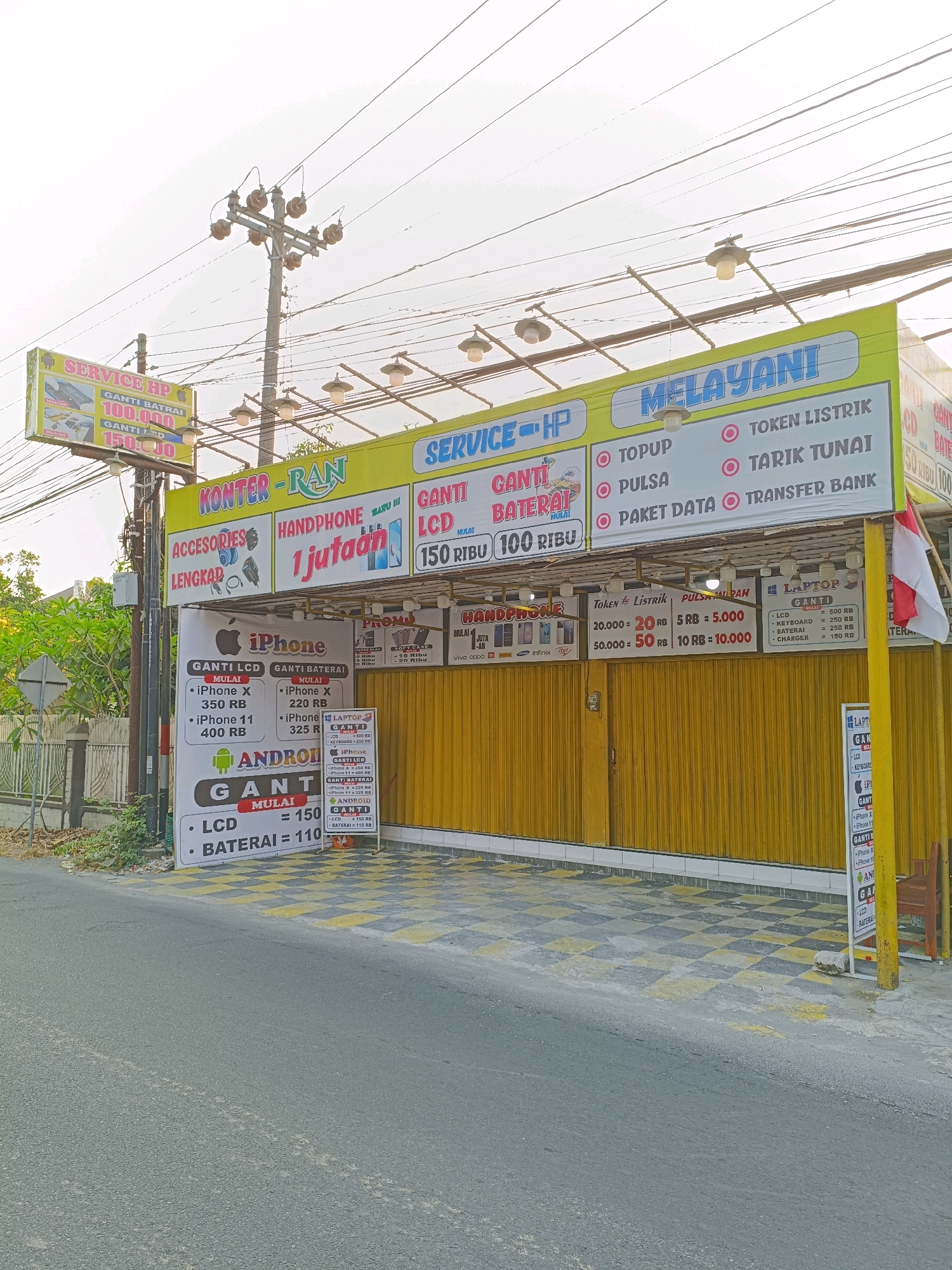 The image depicts a storefront for "KONTER-RAN" and "SERVICE-HP" in Purwomartani, Indonesia. The establishment appears to be a mobile phone repair and sales shop, offering services such as battery replacement, LCD repair, and selling mobile phones and accessories.
The storefront is brightly lit with a yellow and green awning. Various signs are displayed, detailing services and prices. One prominent sign reads "SERVICE-HP MELAYANI" followed by a list of services including "TOKEN LISTRIK", "TOPUP", "PULSA", "TARIK TUNAI", "PAKET DATA", and "TRANSFER BANK". Another sign advertises "GANTI BATRAI 100.000" and "GANTI LCD 150.000".
Below the awning, there are more detailed price lists for different phone models and services. For example, there are listings for iPhone repairs (GANTI LCD, GANTI BATERAI) and Android repairs. The shop also seems to sell accessories, indicated by a sign with pictures of headphones and other items labeled "ACCESORIES LENGKAP".
The exterior of the shop features a yellow roll-up door. A small white sign with a red and blue logo that appears to be related to mobile services is placed near the entrance. A lamppost with multiple lamps is mounted above the awning.
The environment surrounding the shop includes a sidewalk with a black and yellow checkered pattern. A street is visible in the foreground, and some greenery and a wall are on the left side of the image. Overhead, numerous electrical wires crisscross the sky, typical of many urban and semi-urban areas in Indonesia. No people are clearly visible in the image. The overall impression is that of a functional, local business operating in a well-trafficked area.