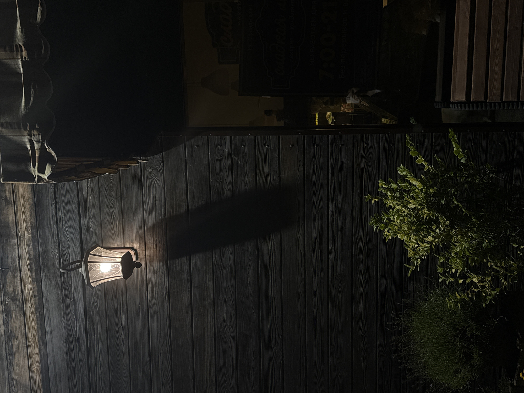 The image shows the exterior of a building at night, likely a shop or cafe, in Abinsk, Russia. A vintage-style lantern with a glowing bulb is mounted on a dark wooden wall, casting a long shadow. To the right, an awning shades a window, through which some interior lights and signage are visible. A dark sign on the window appears to advertise "сладкая и" which translates to "sweet and", followed by "пекарня кондитерская" meaning "bakery confectionery". Below this, text indicates operating hours: "МЫ РАБОТАЕМ ДЛЯ ВАС" (We work for you) and "7:00 - 21:00". Two green bushes are visible in the foreground, one on the left and another on the right. The overall scene is dimly lit, with the primary light source being the lantern and the interior of the building.