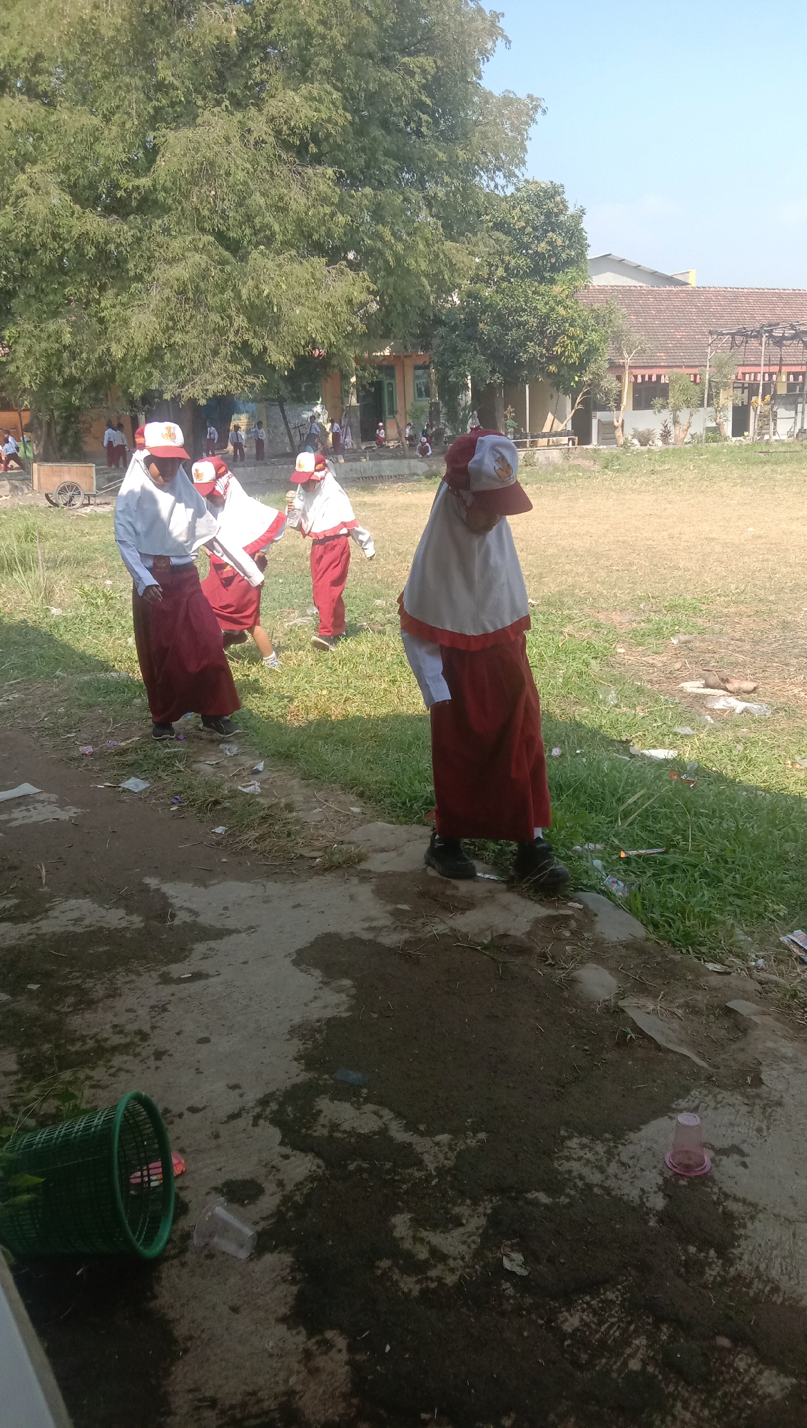 In Karanganyar, Indonesia, a group of young students in school uniforms are gathered in an outdoor area. The students, wearing red skirts and white tops with red hats and white headscarves, appear to be engaged in an activity on a grassy field. The environment includes trees, a building in the background, and a paved area with some litter, including a green trash can and plastic cups. The sun is shining, indicating it is daytime.